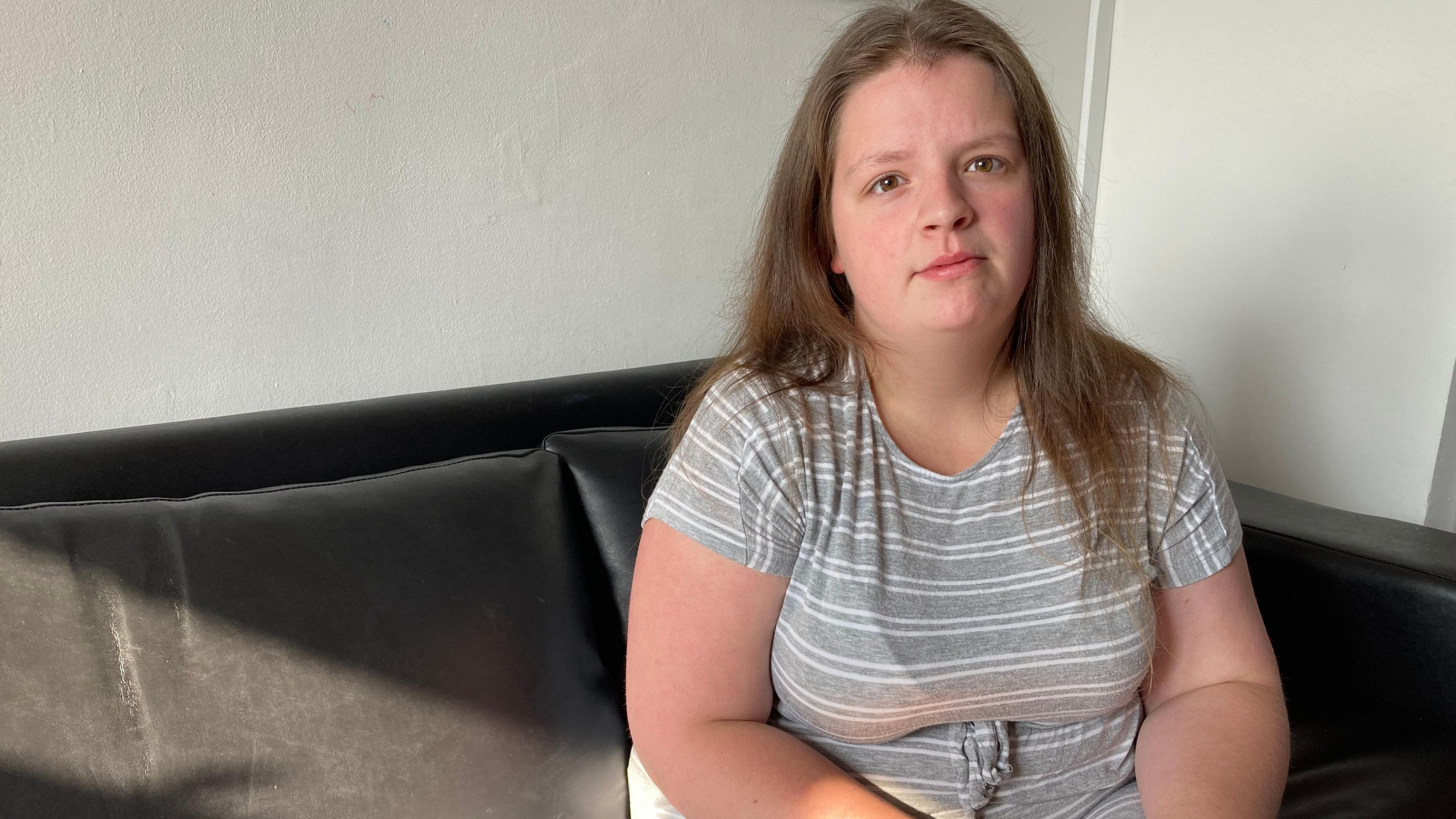 Aimee Richards, who has light brown hair, sits on a black sofa looking into the camera 