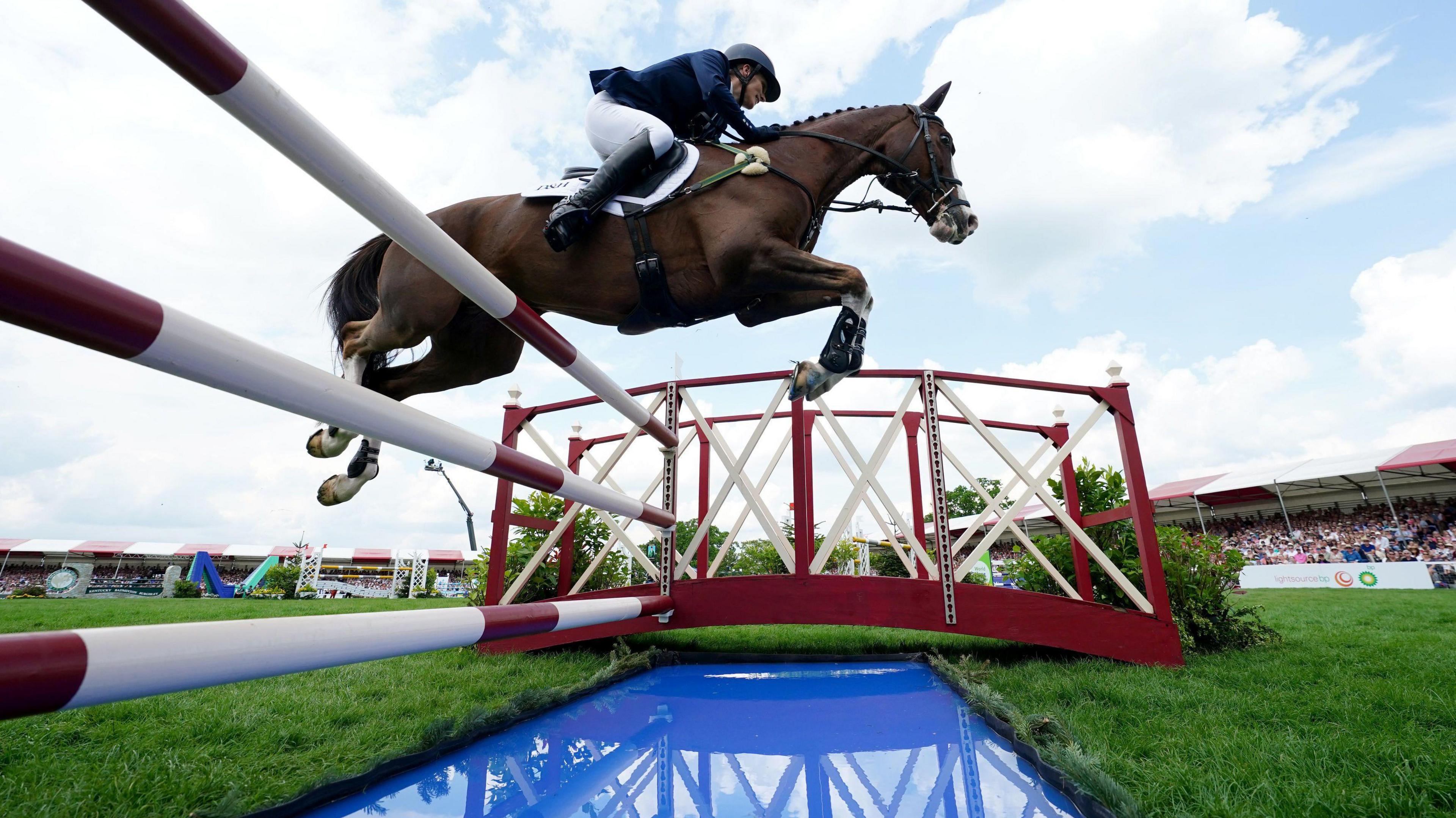 Zara in mid flight on her horse over a show jump with red and white poles and a water trough.