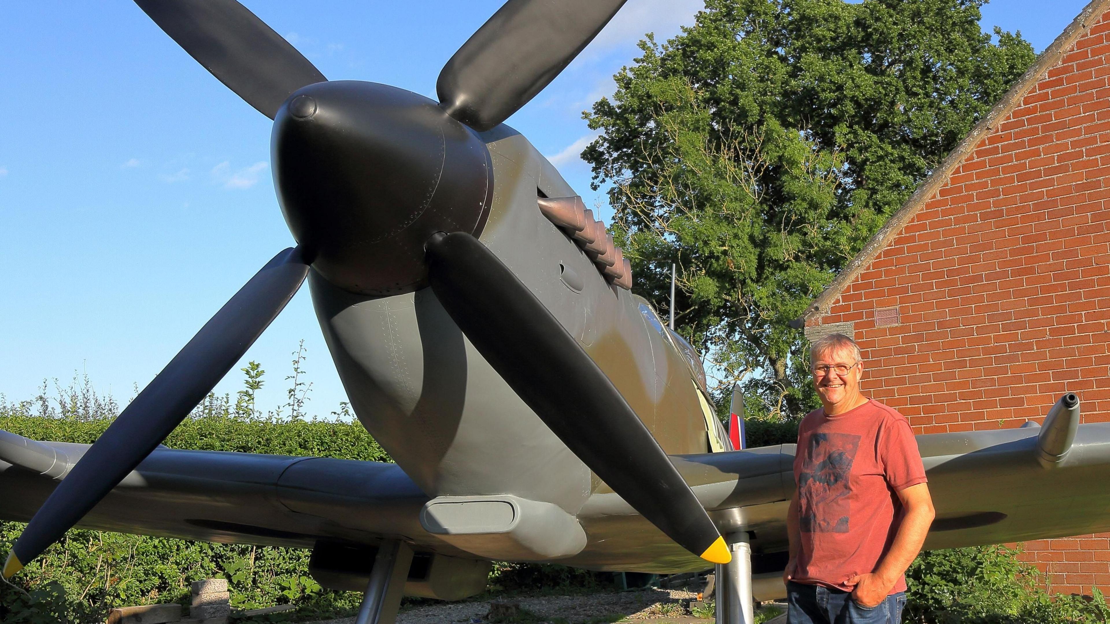 David Price standing next to his spitfire. He has short white hair and glasses and is wearing a red t-shirt. Behind him is a red-brick gable end of a house. The spitfire is painted a brown-grey colour.