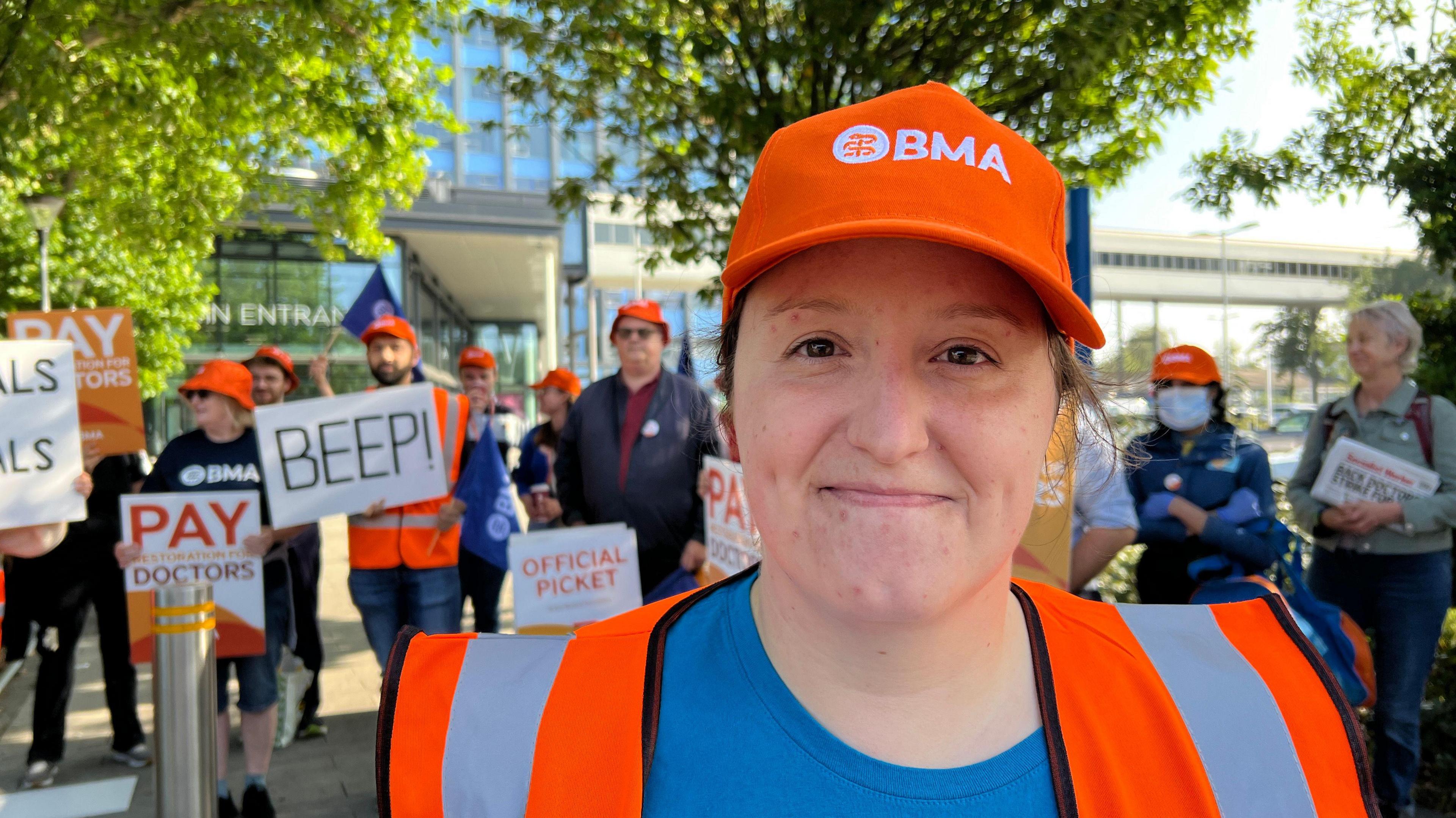 A woman with her hair tied back wearing a orange BMA hat and orange hi-vis top with a blue top underneath. She is smiling at the camera. Behind her are several people holding up signs.