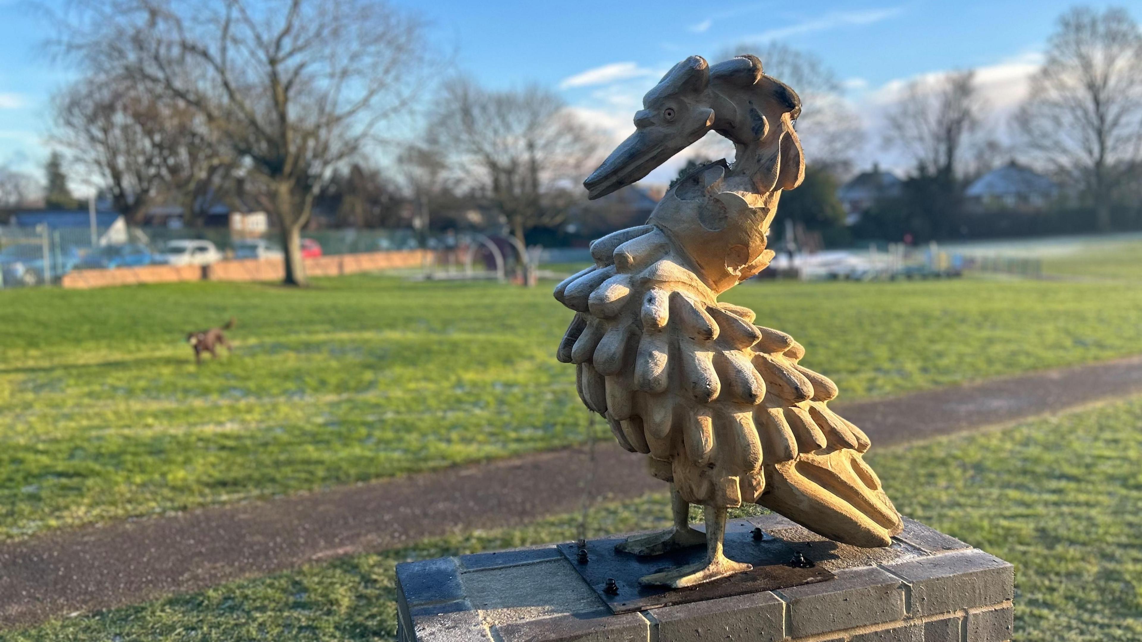 A wooden swan statue with a crown around its neck which has a small chain attached to it linked to a plinth on top of a brick base. The backdrop is a park with a recreation area in it. 