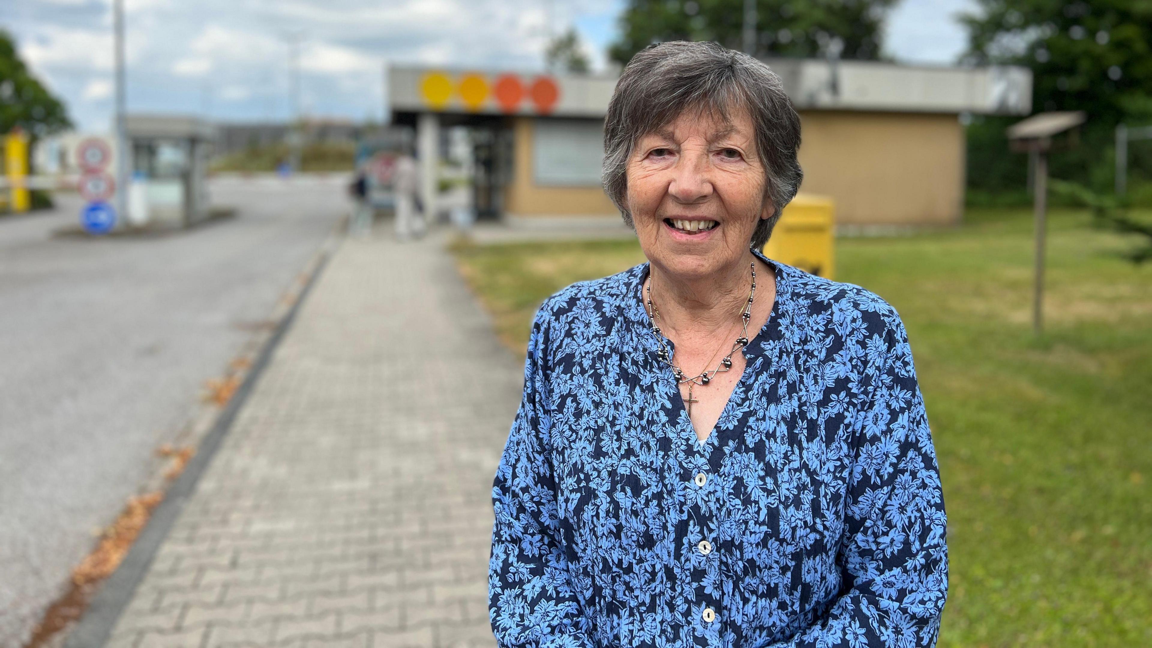 Sylvia stands on a paved pathway next to a grassy area. They are wearing a blue floral-patterned shirt and a necklace. In the background, there is a small building with colorful circular signs on its roof, along with trees and a cloudy sky