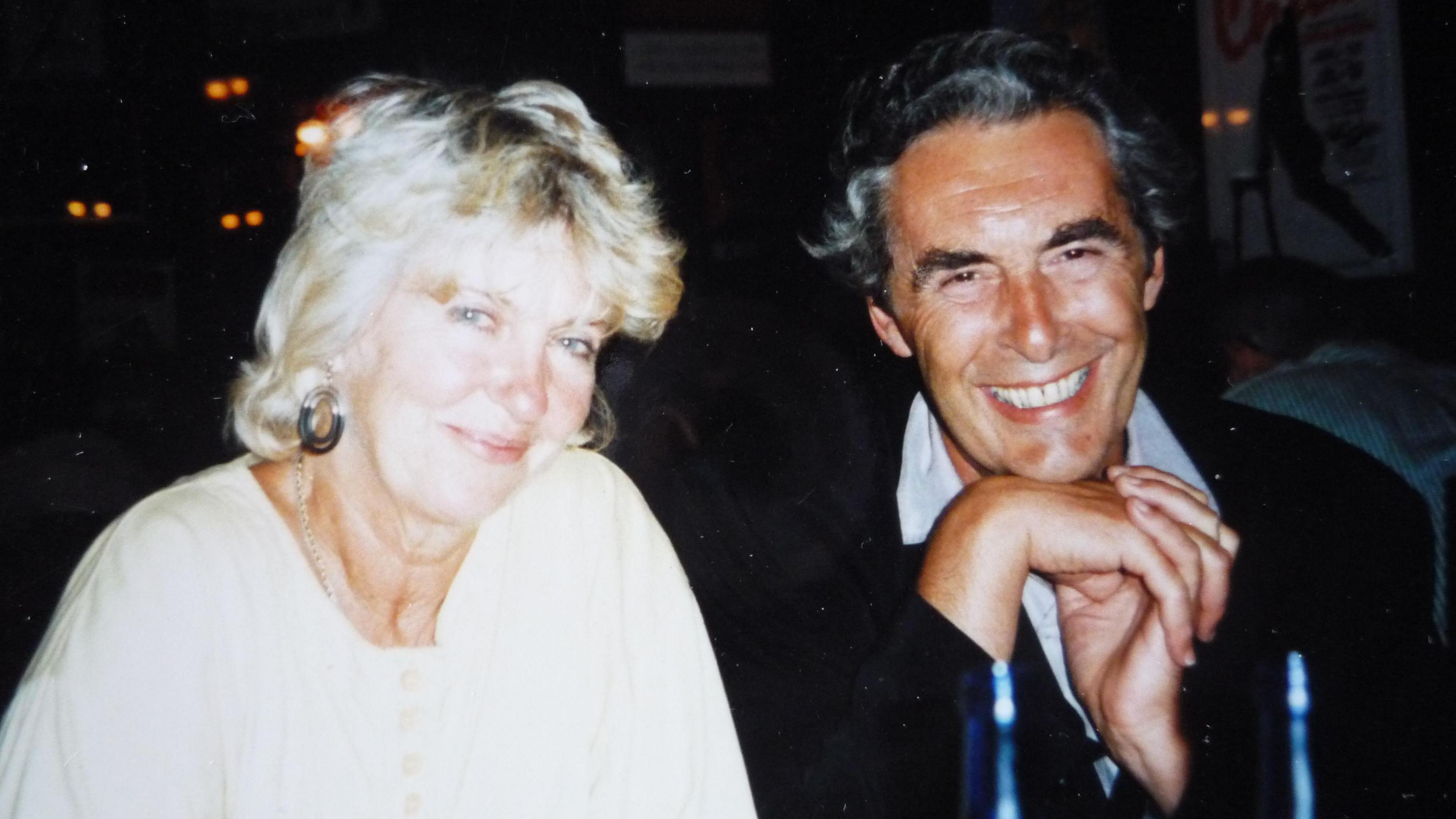 Tony Mathews smiles as sits with his hands clasped under his chin, wearing a dark jacket and white shirt while sitting to the right of his smiling wife Narissa Mathews who is wearing a cream blouse with cream buttons. 