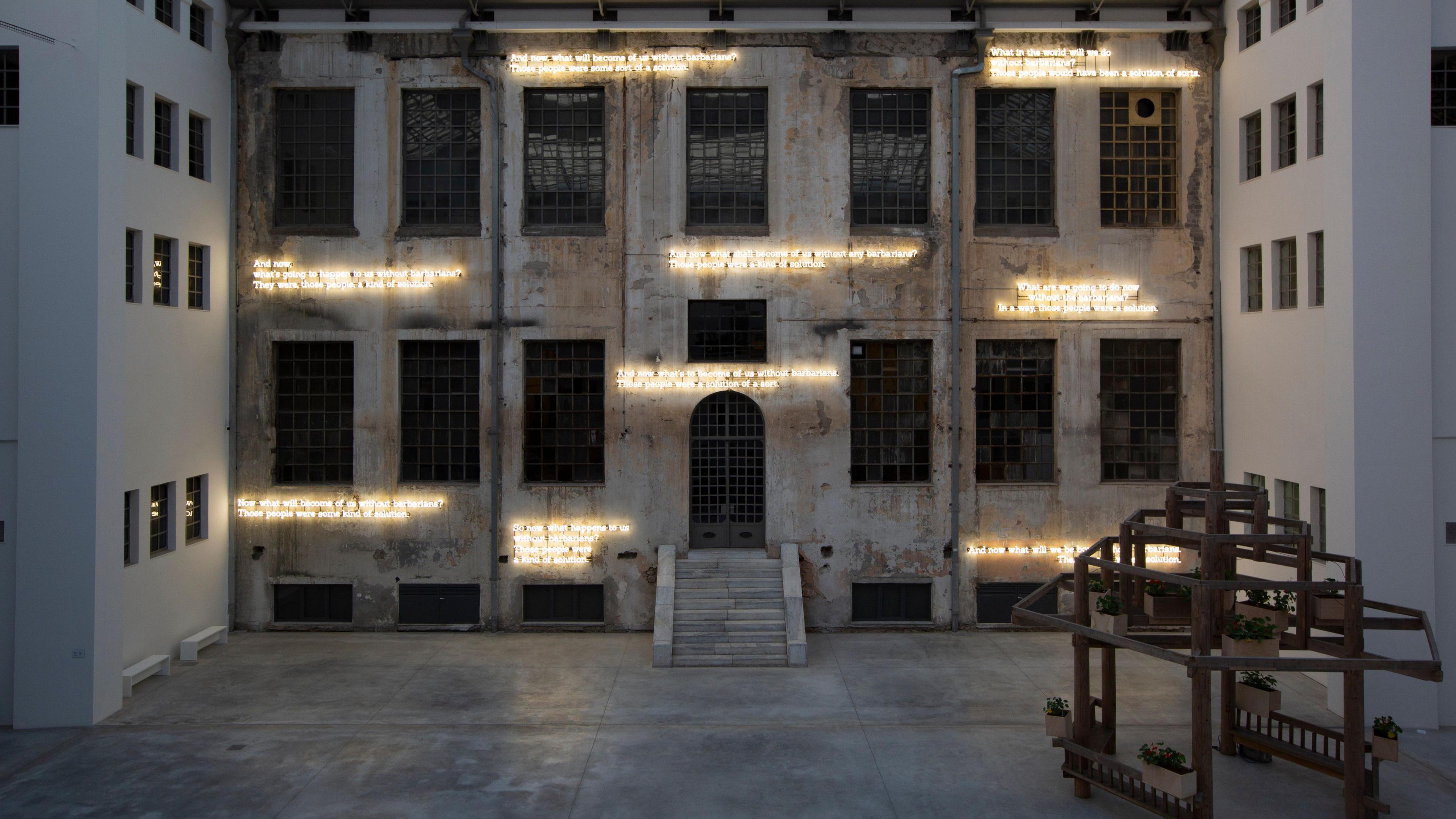 A grand industrial-looking white building with a stone staircase up to the front door. Dotted around the facade are neon lights spelling out various words.