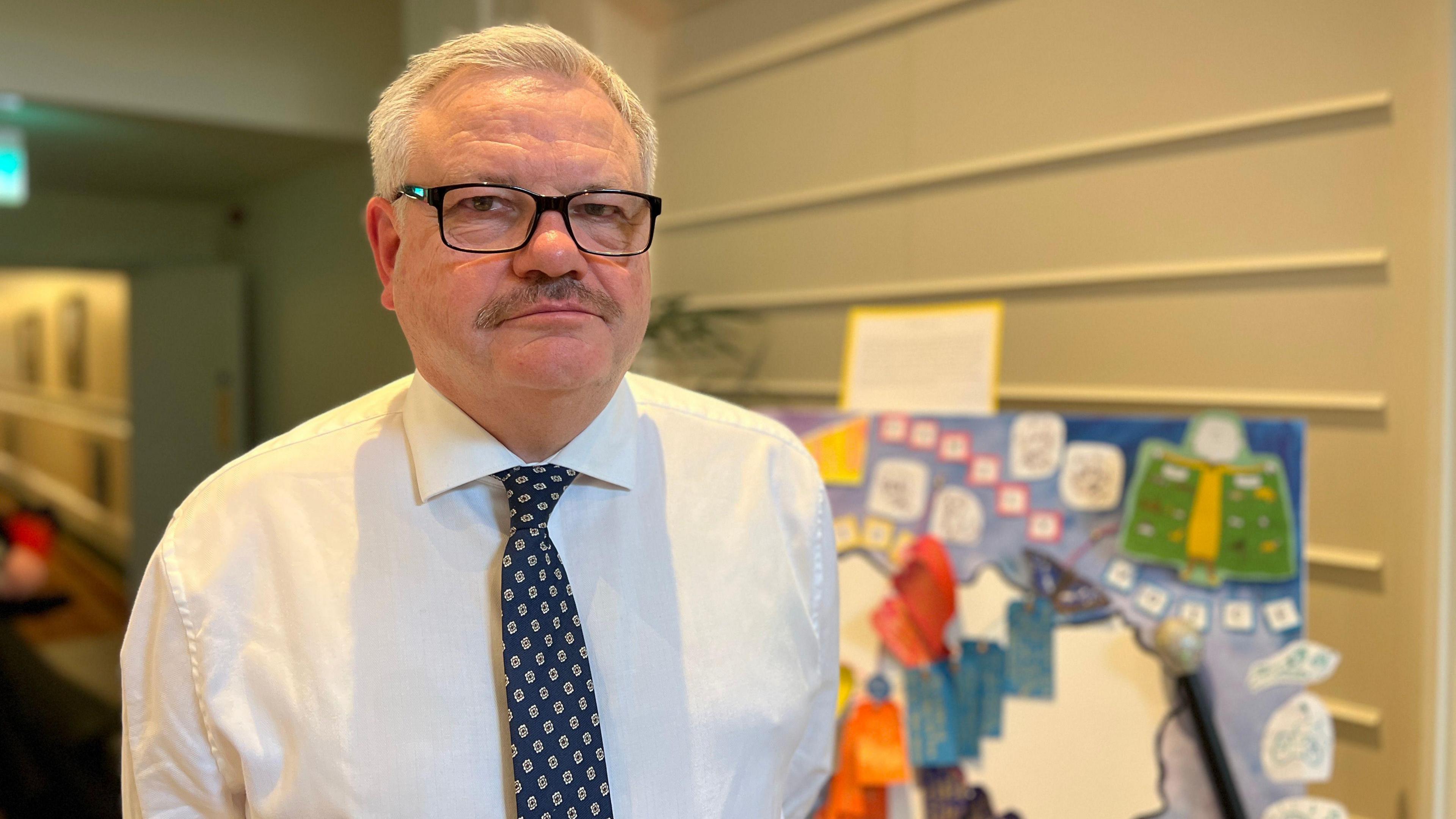 Richard looks at the camera with a piece of art made by the children in the background to his right. He's wearing a white shirt with a blue tie which has patterns on. 