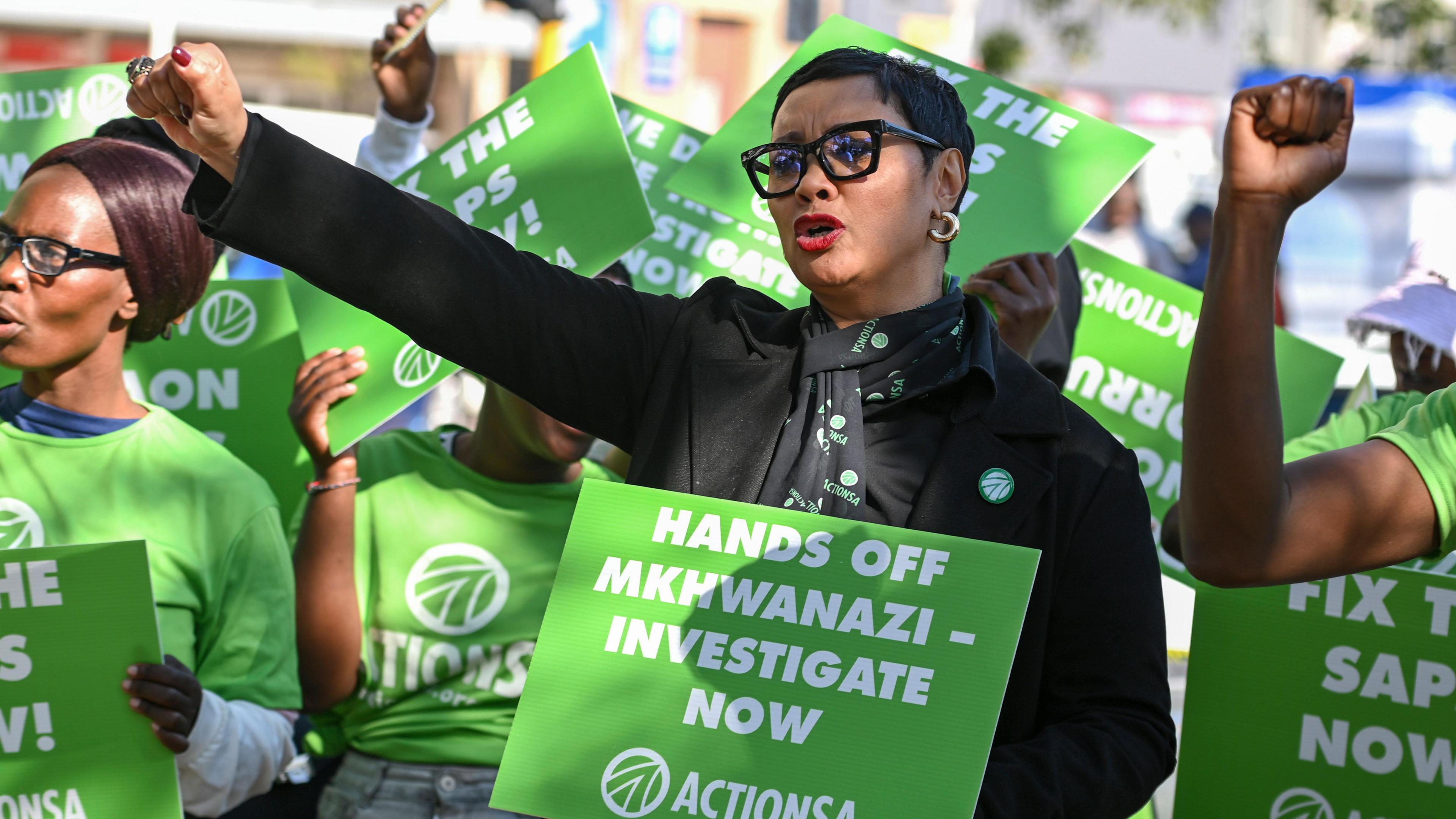Protesters holding placards reading: Hands off Mkhwanazi - Investigate now. One woman wearing a black shirt has her first raised, others are wearing green T-shirts