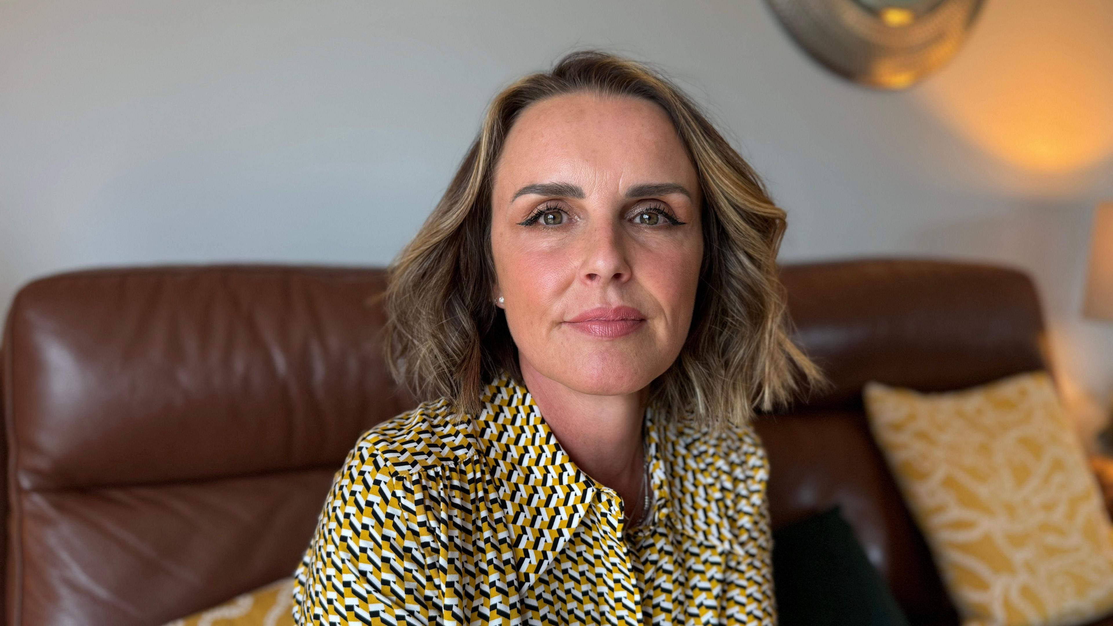 Rachel, who has blonde-brown hair, looks at the camera. She is sat on a brown leather sofa that is scattered with yellow and white cushions. She is wearing a black, white and yellow shirt.