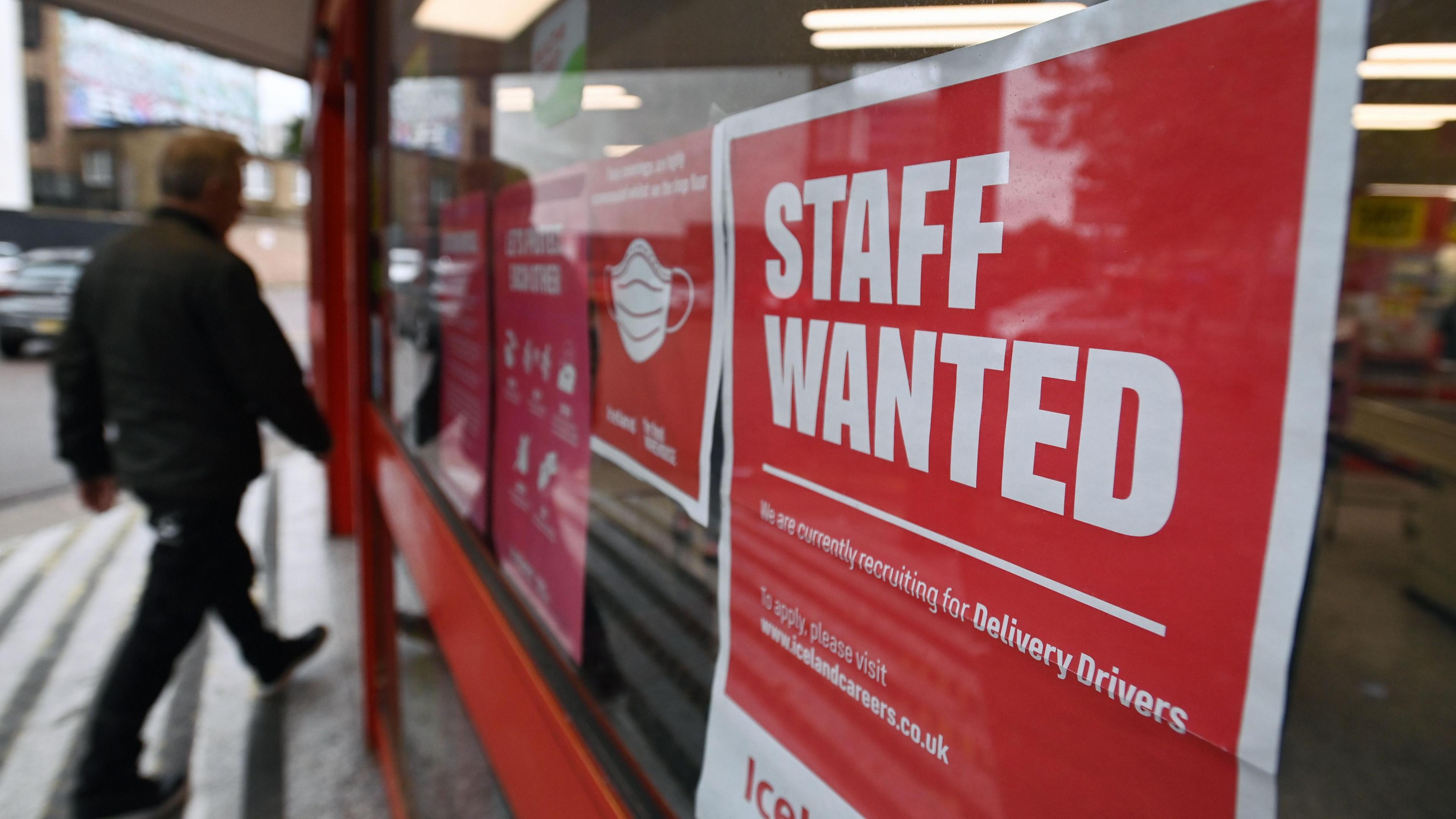 A man walks past a sign in a shop window with the words staff wanted in white letters on a red background