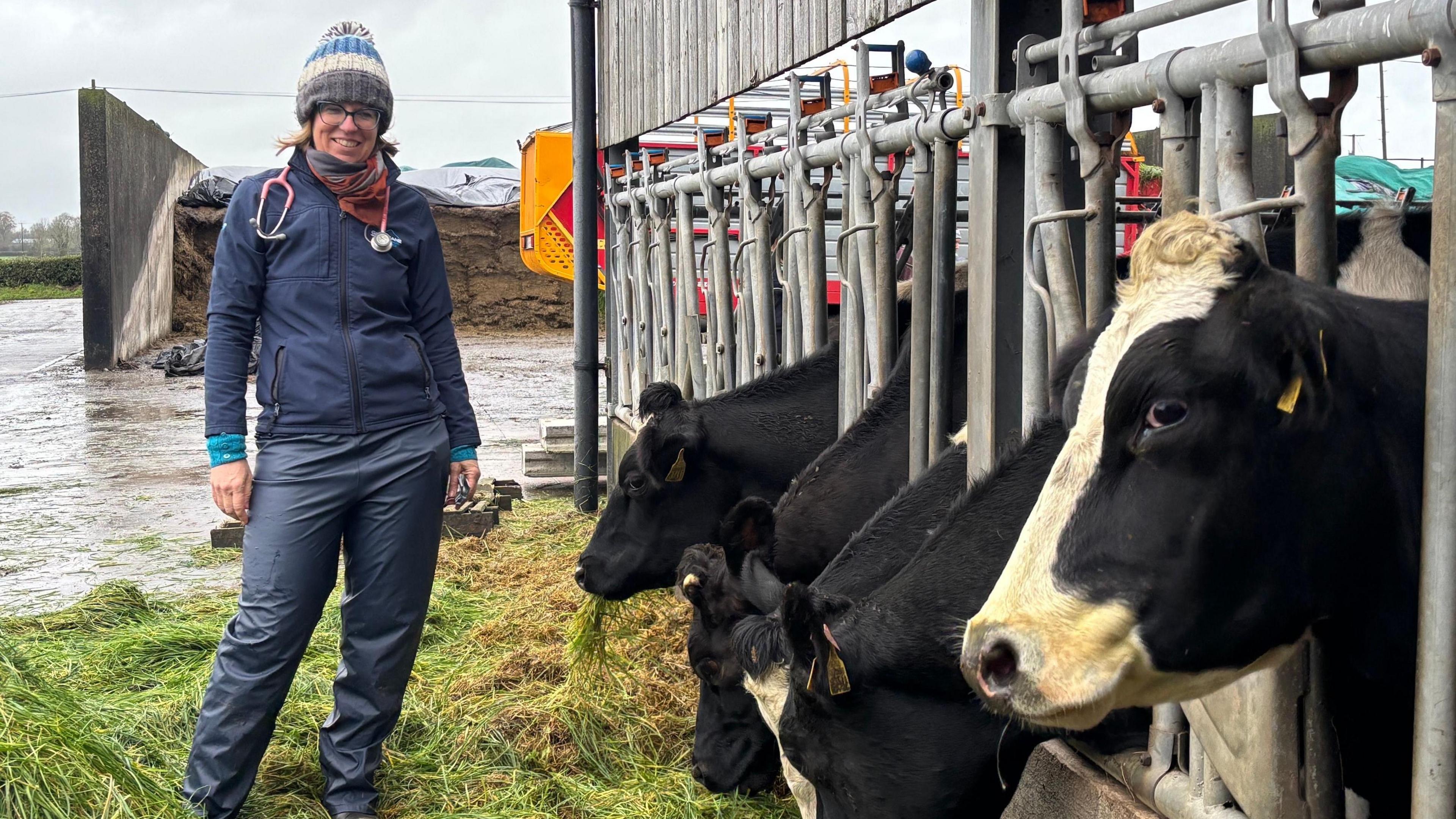 A vet stands in a farmyard. She is dressed in a navy top and waterproof trousers with a colourful bobble hat. She has a stethoscope around her neck and a wide smile. Five cow heads poke through the bars of their shed to reach some grass on the ground. The dairy cow biggest in frame is side-eying the camera.