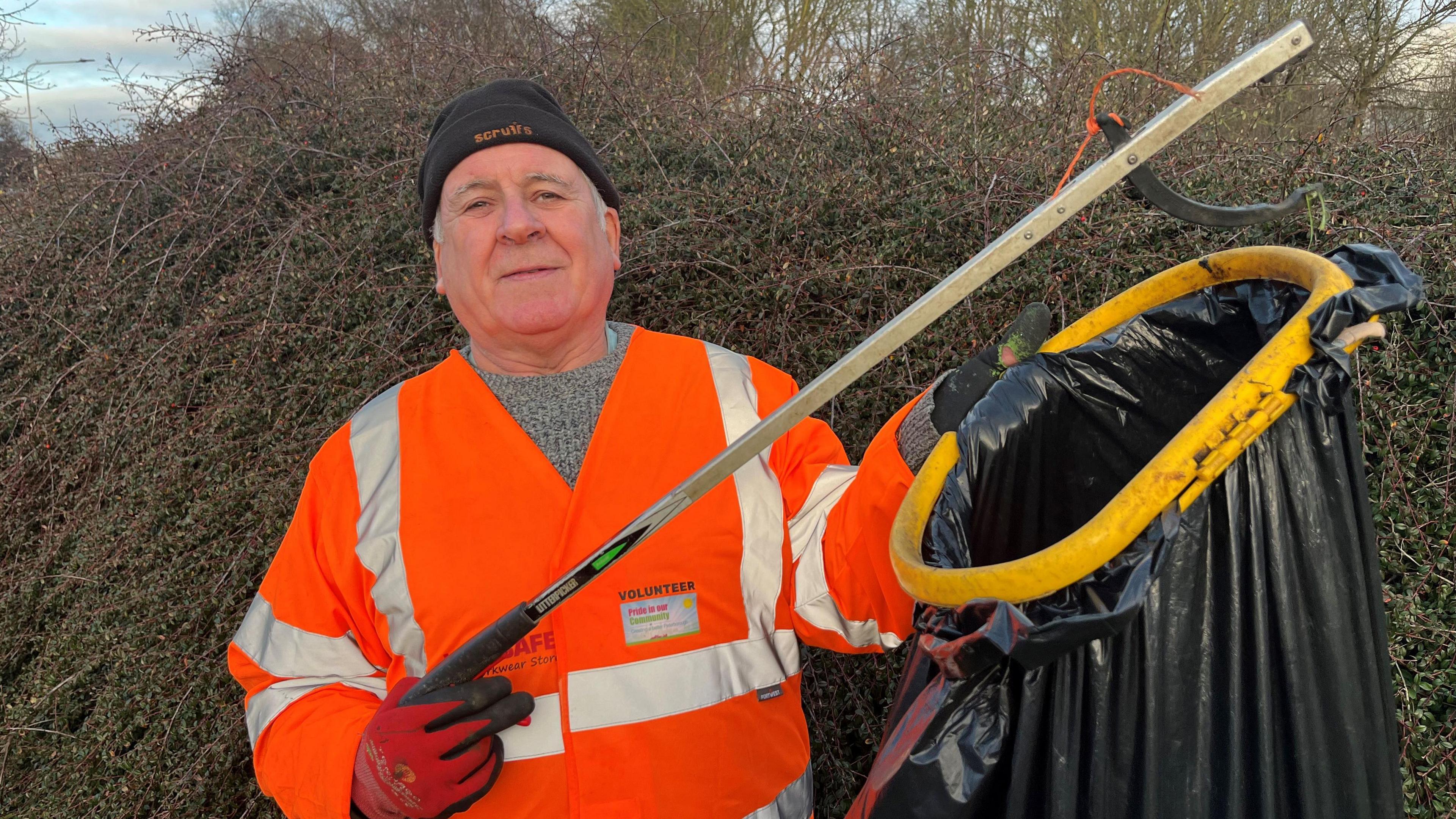 Mark Fishpool, 68, has a black beanie hat on with some grey hair and sideburns visible. He is wearing a hi-viz orange jacket with a grey jumper underneath, and red gloves. He is holding a litter grabber in one hand and a black bin liner in the other and there is a hedge behind him,