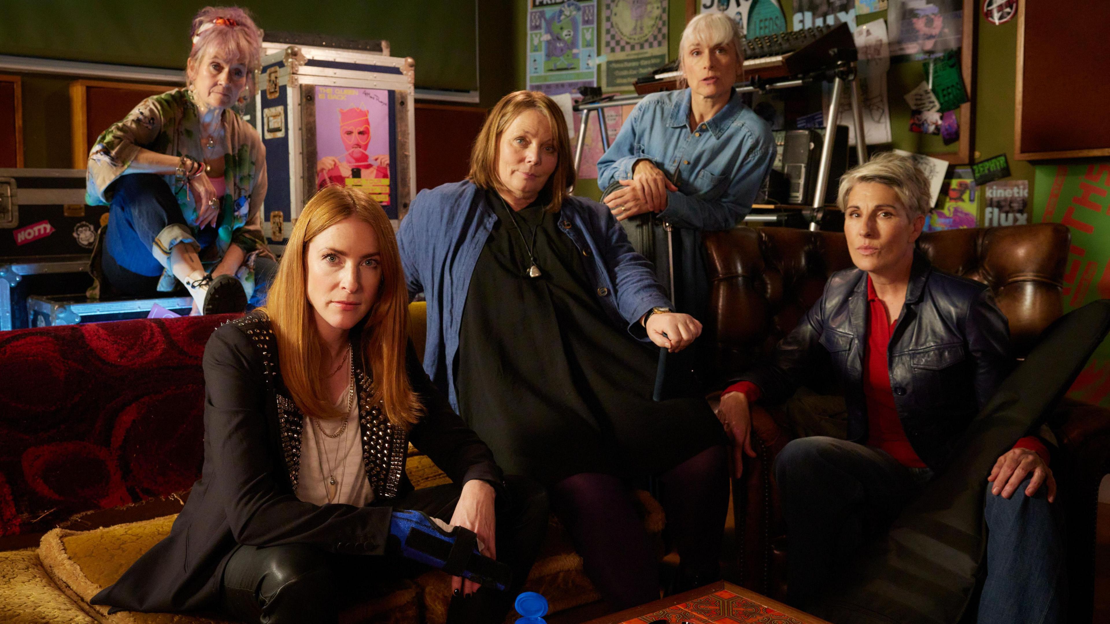 The image depicts five individuals gathered in a relaxed, creative indoor setting. They are positioned around a coffee table that holds a blue cup, a water bottle, and some scattered papers. The room is decorated with posters and artwork on the walls, suggesting an artistic or musical theme. The people are dressed casually and appear to be either engaged in conversation or posing for the photograph. The furniture includes sofas and armchairs, contributing to the informal and comfortable atmosphere of the space.