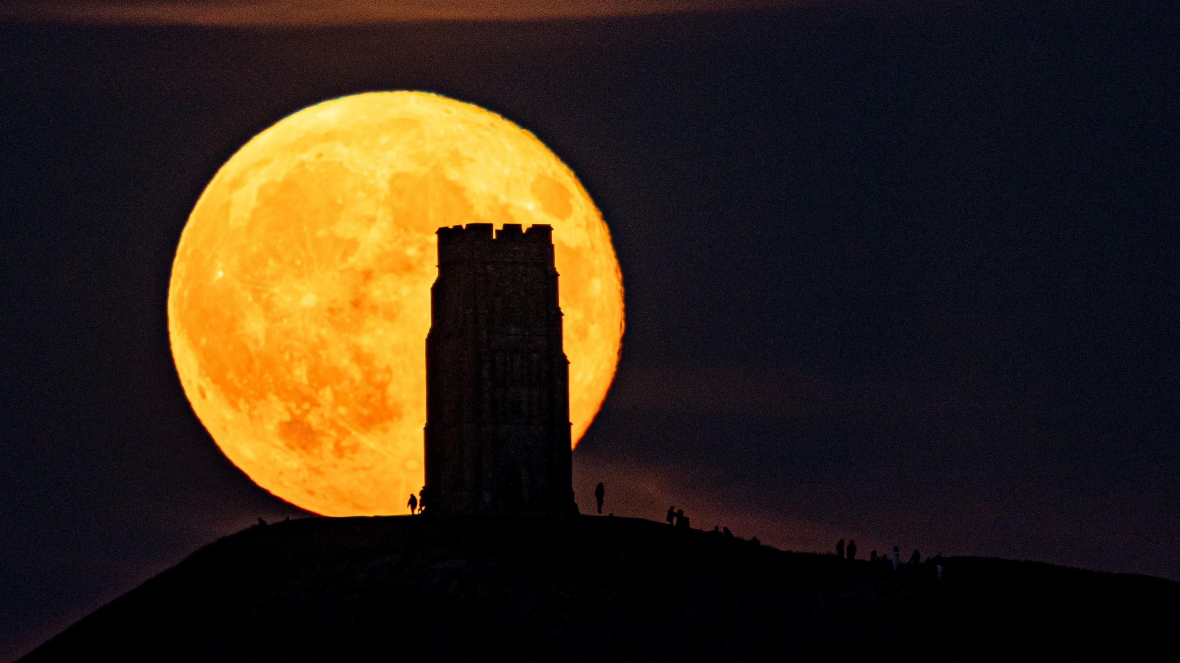 The full moon, large and bright, orange in colour rises behind Glastonbury Tor with people standing in shadow