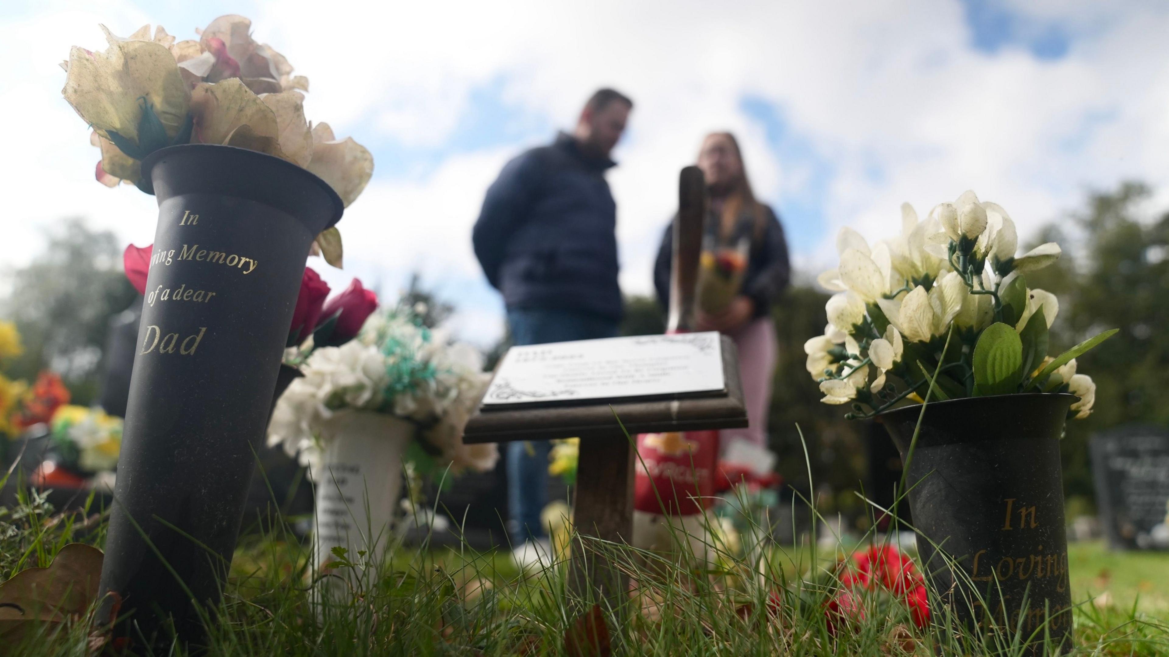 In the foreground of the picture are several pots of flowers, one with the inscription "In loving memory of a dear dad". In the background a man and woman stand with a bouquet of flowers.