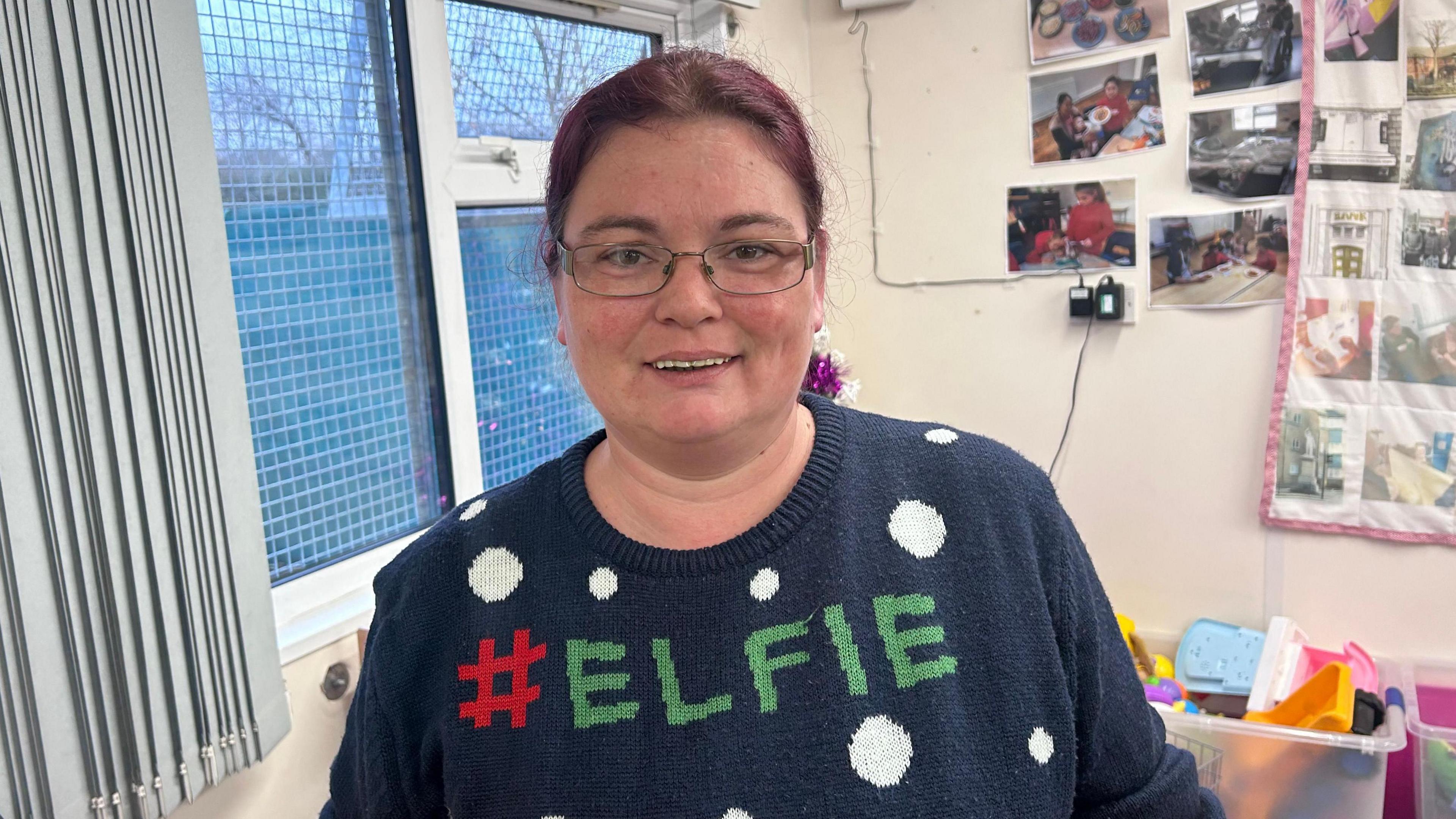 Victoria Barlett smiles into the camera. She has plum-coloured hair and is wearing glasses. She is wearing a dark blue Christmas jumper which features white spots and has the word "Elfie" written in large capital letters. She is stood in a white room and toys and photographs are visible in the background.