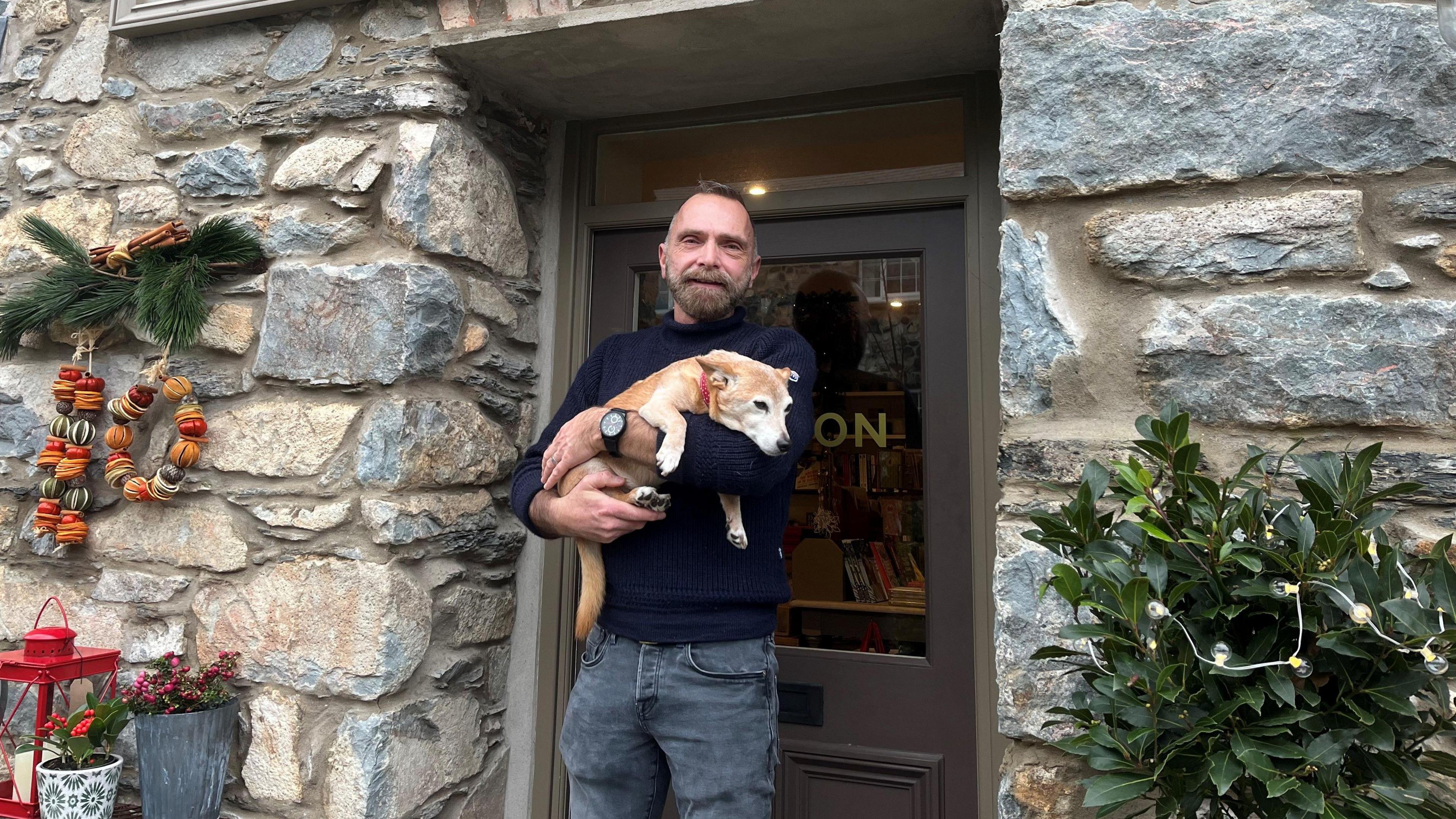 Tomos Meredith outside a shop with stone walls and some hanging Christmas decorations. He has a beard, short hair and is wearing a blue jumper and grey trousers. He is holding a small dog