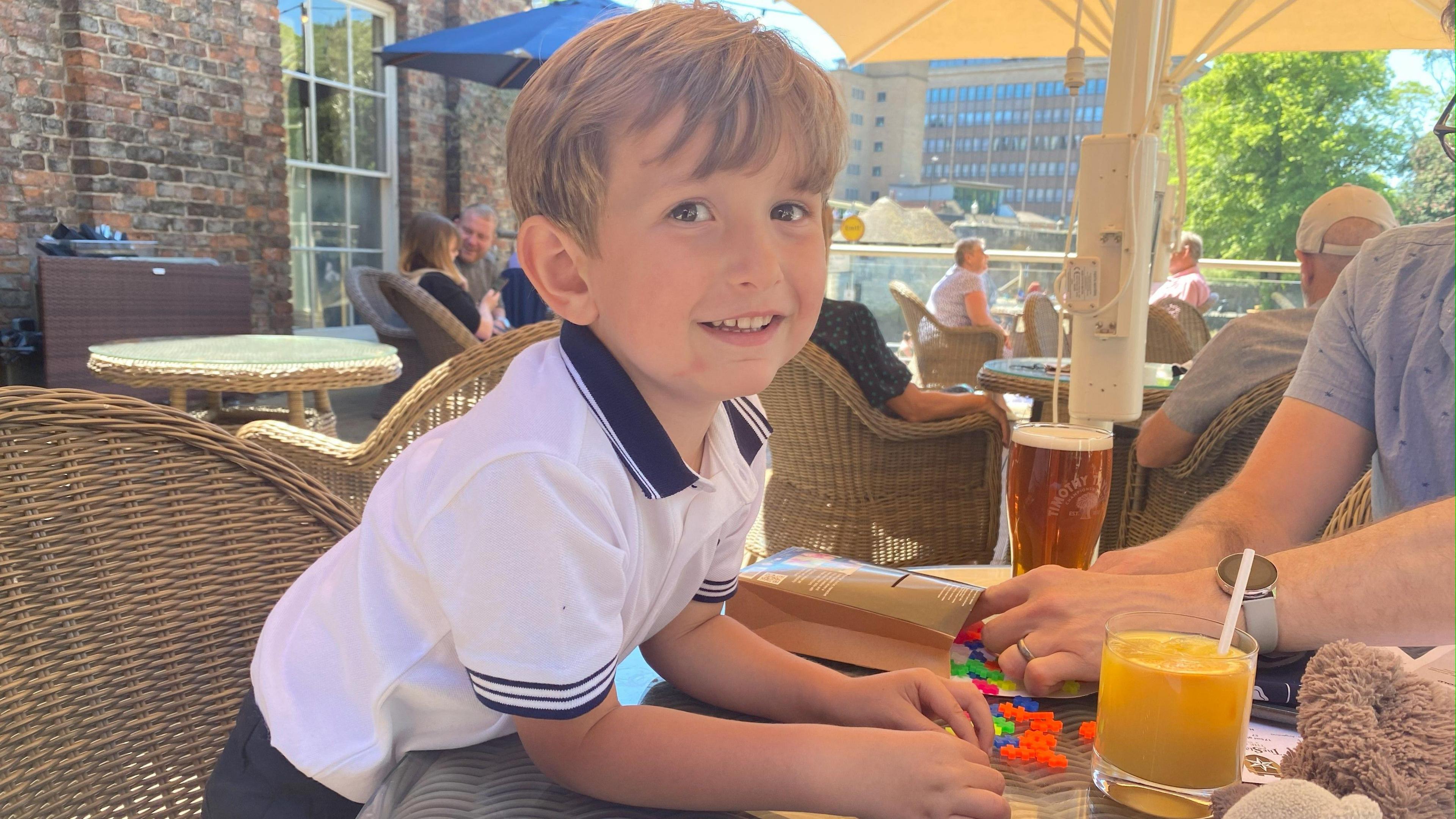 A young boy with dark blond hair with a fringe across his forehead is leaning forward on to a table from a wicker chair and smiling at the camera. He is wearing a white and black polo shirt. On the table is a glass of orange juice and some colourful building blocks that an adult is setting up.