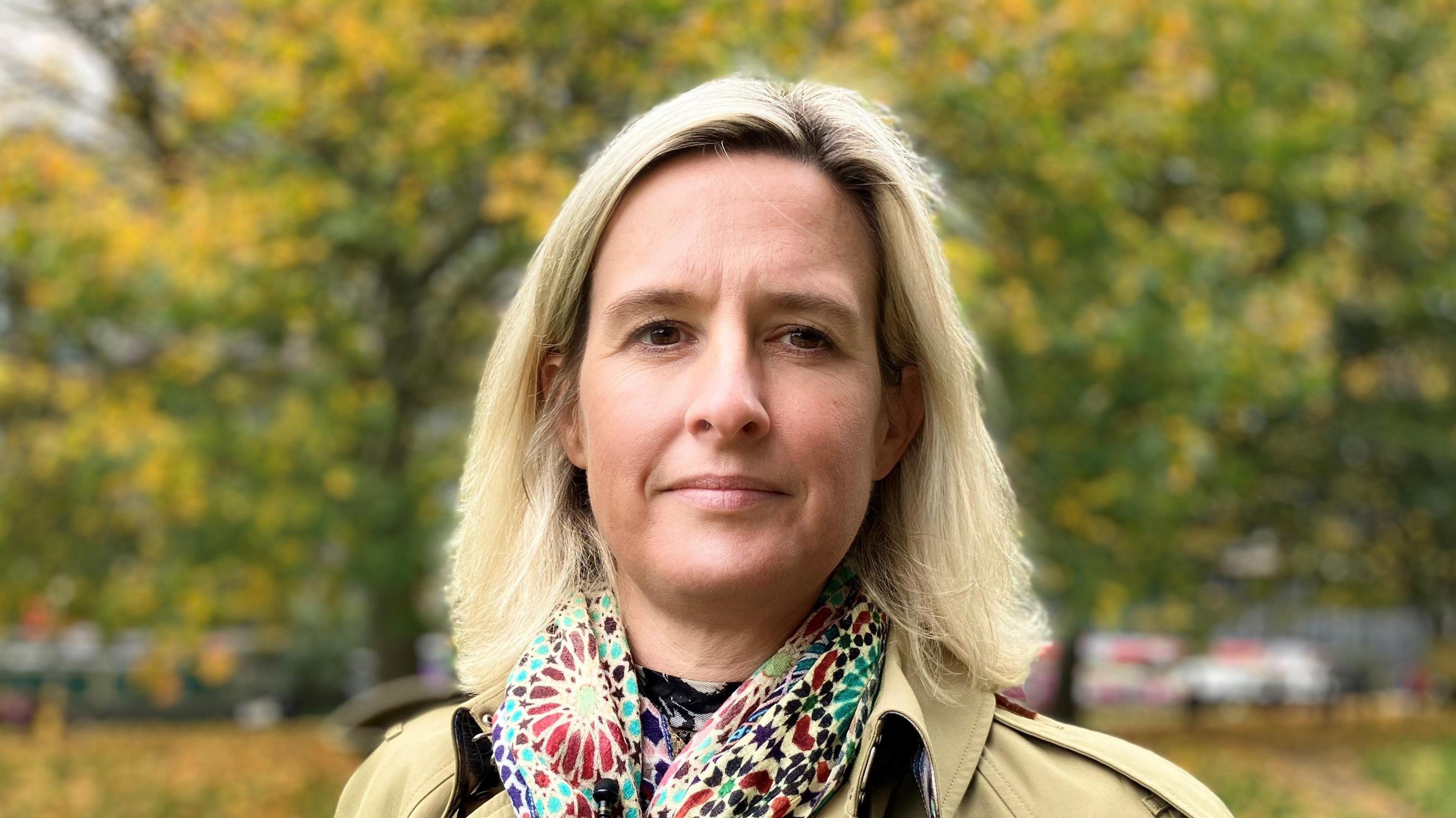 Gemma Lindfield, a woman with short blonde hair wearing a multicoloured patterned scarf and beige trench coat, stood in front of some trees