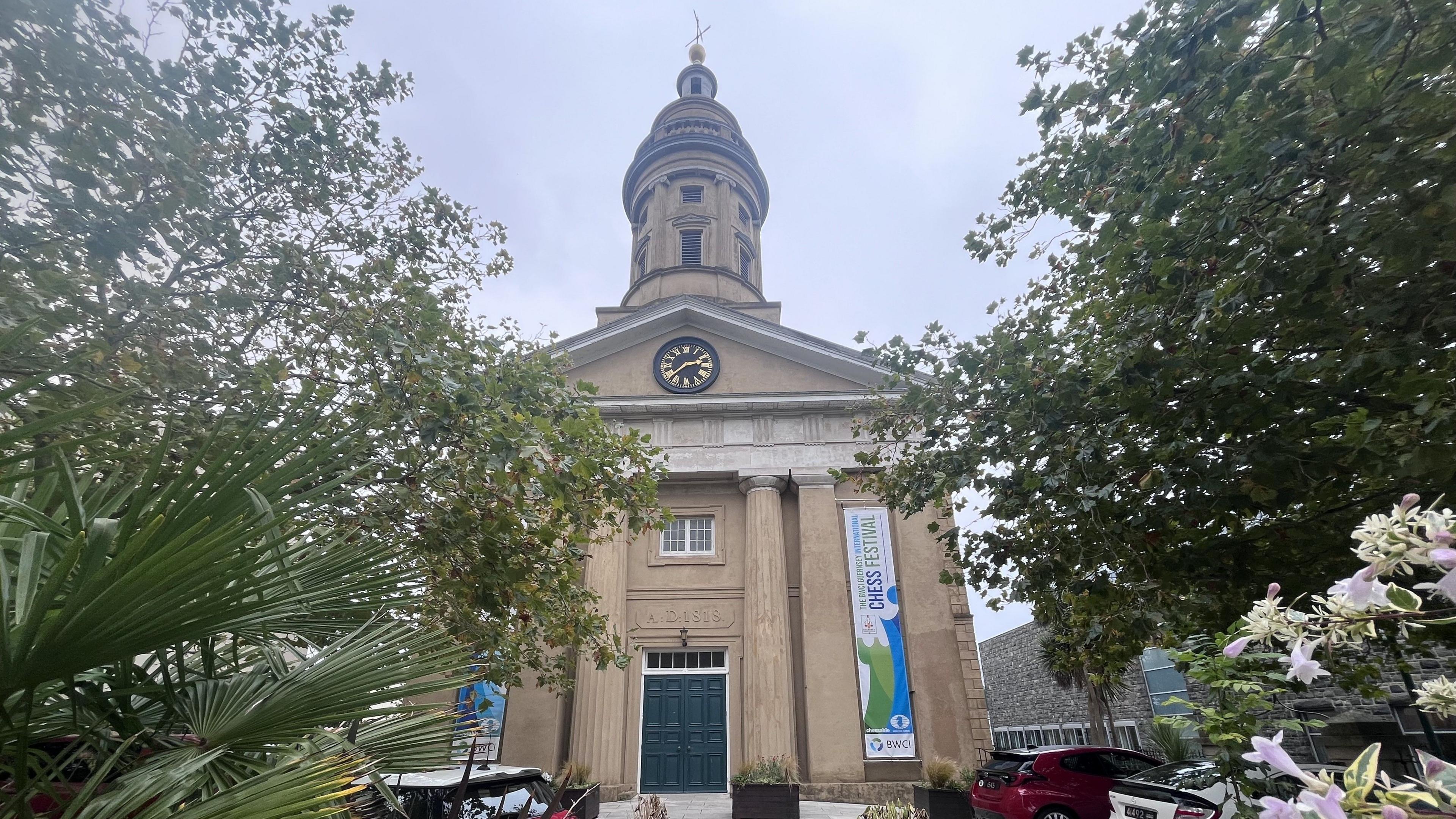 A large converted limestone church building, with Roman-style pillars, a black and gold clock face, and a tower with a gold sculpture on top.