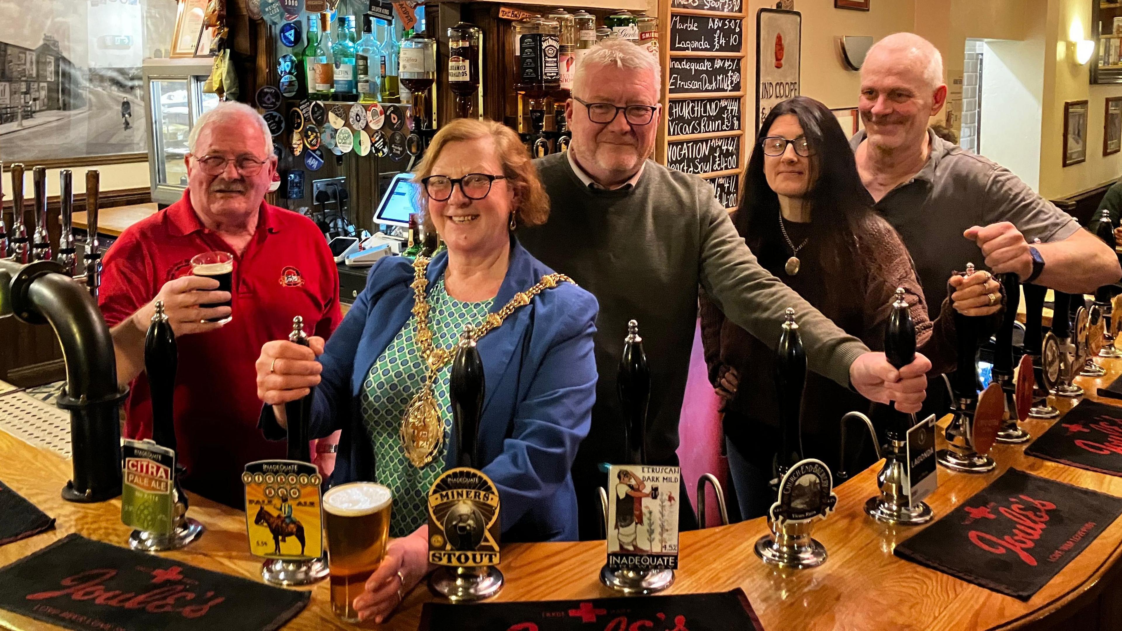 Two women and three men standing behind a bar at a pub. Some are holding the beer pumps while others are holding drinks. One of the women is wearing a mayoral chain.