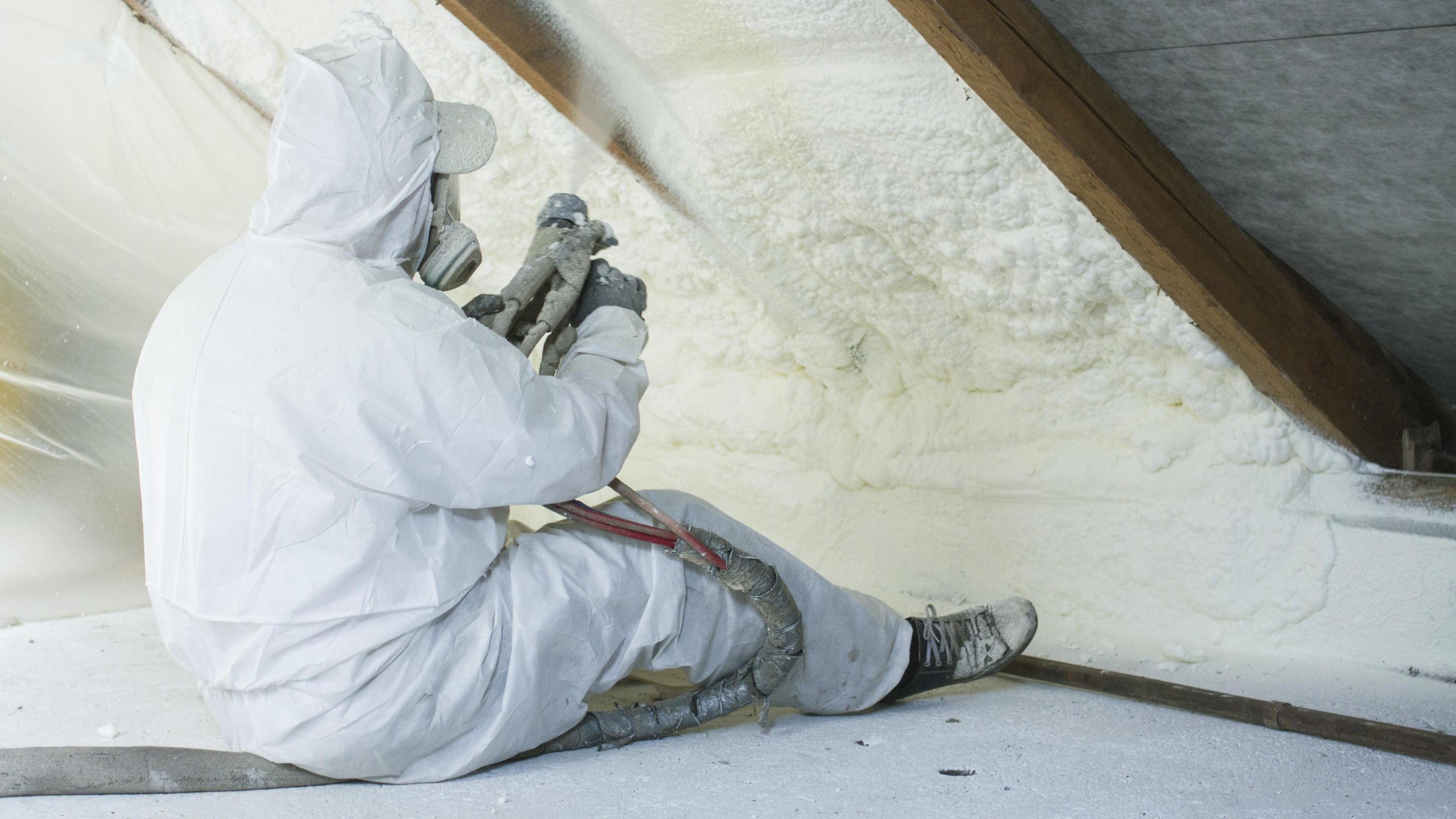 Tradesman sits on the floor of a loft space, using tools to apply spray foam insulation to the underside of the roof while wearing a white set of overalls and a respiratory mask