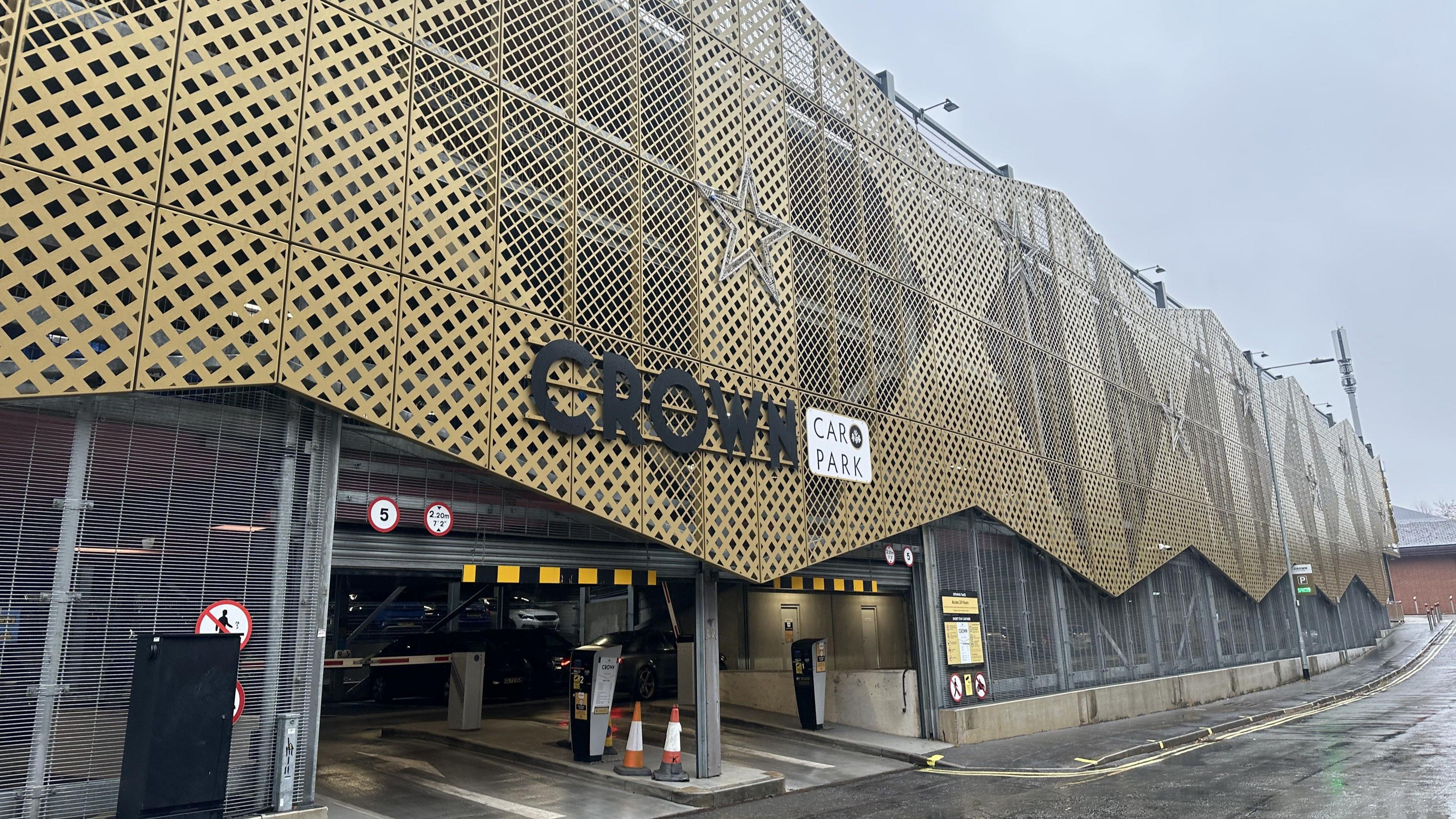The image shows the exterior of a multi-story car park named Crown Car Park. The building features a distinctive architectural design with a facade made of gold-coloured lattice panels arranged in a geometric pattern, creating a textured and modern appearance. The panels are mounted on a metal framework that curves slightly, giving the structure a dynamic look.
At the entrance, there is a large black sign with bold letters spelling CROWN, accompanied by a smaller white sign that reads CAR PARK. The entrance is wide enough for two lanes, with yellow and black clearance bars overhead indicating height restrictions. Several traffic signs are visible, including speed limits and warnings, and two orange-and-white traffic cones are placed near the entry point. Inside the entrance, a dark-coloured vehicle is partially visible, along with ticket machines and barriers for controlled access.