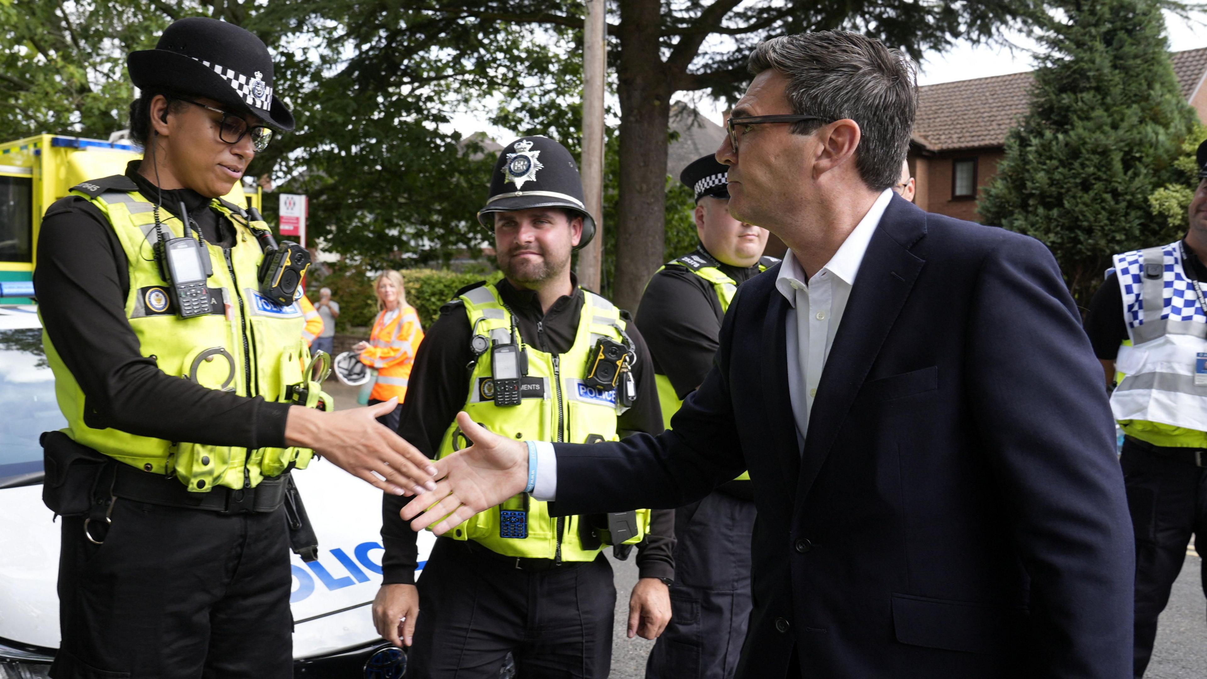 Andy Burnham, a man with dark hair, black glasses, and wearing a dark coloured blazer and white shirt, shakes hands with a police officer in uniform while three others look on. A police car can be seen in the background, as well as houses and trees.