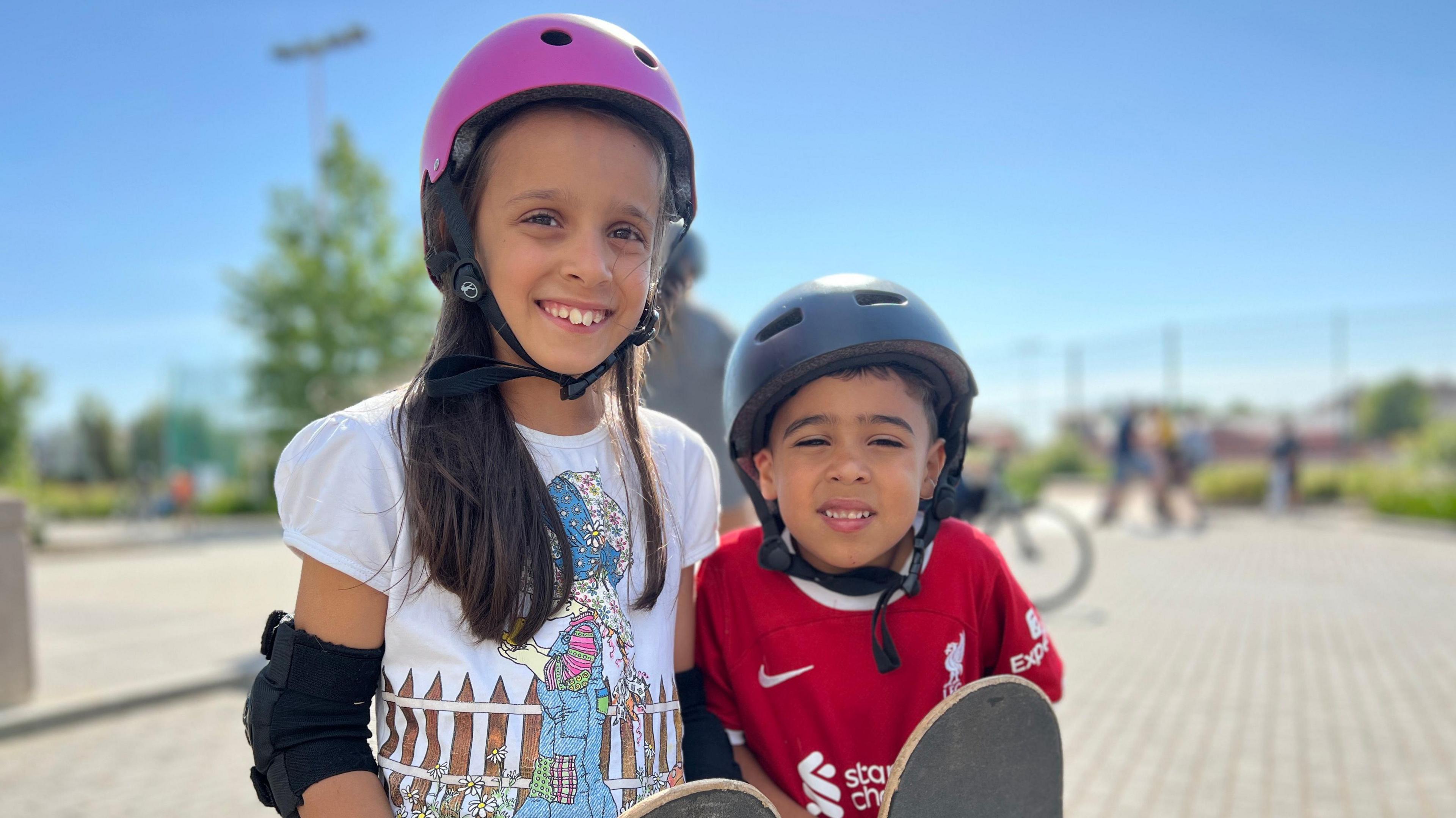 Melissa and Theo smile at the camera as they wear helmets and elbow pads. Melissa has long brown hair which comes past her shoulders and is wearing a white t-shirt with a cartoon girl by a fence on it and Theo is wearing a red Liverpool FC top. They're both holding skateboards.