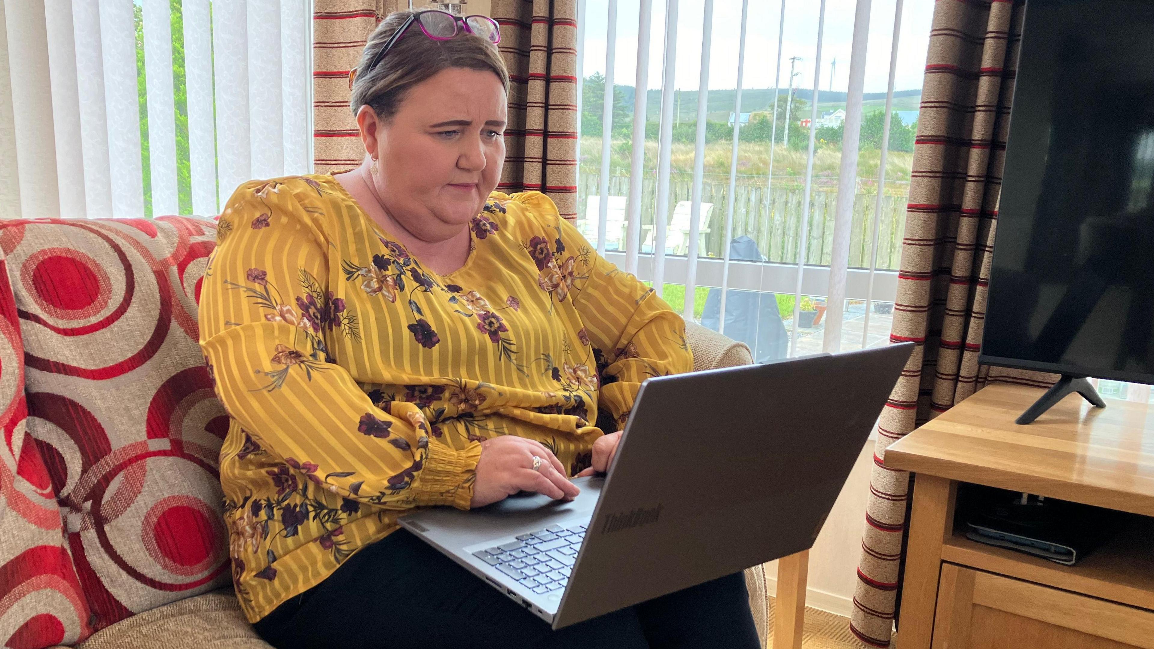 Joy wearing yellow top with floral pattern sitting on a sofa with an open laptop in her mobile home