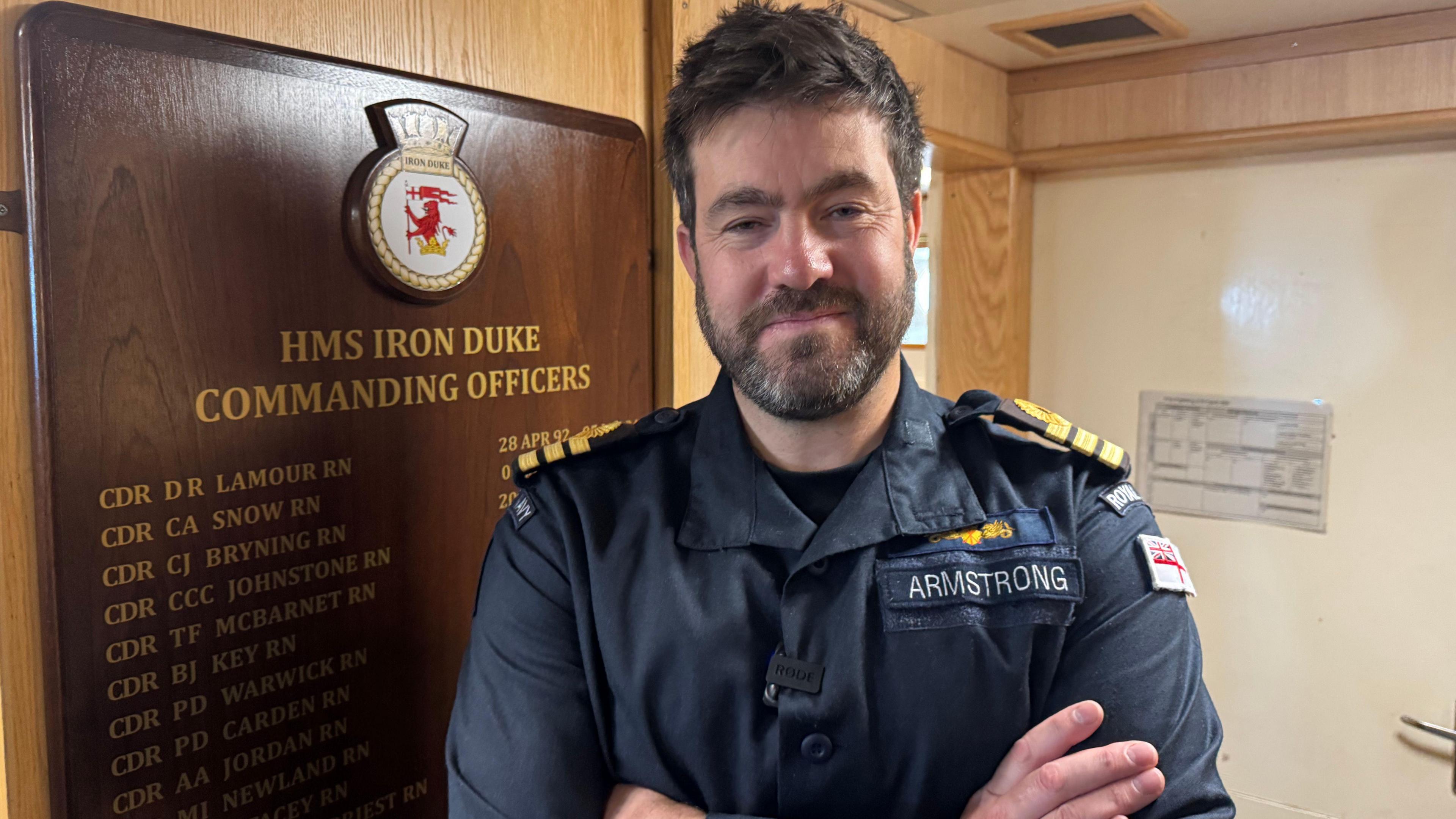 The captain of HMS Iron Duke stands with his arms folded in front a wooden plaque displaying the names of former commanders. He is wearing a blue coloured naval uniform.
