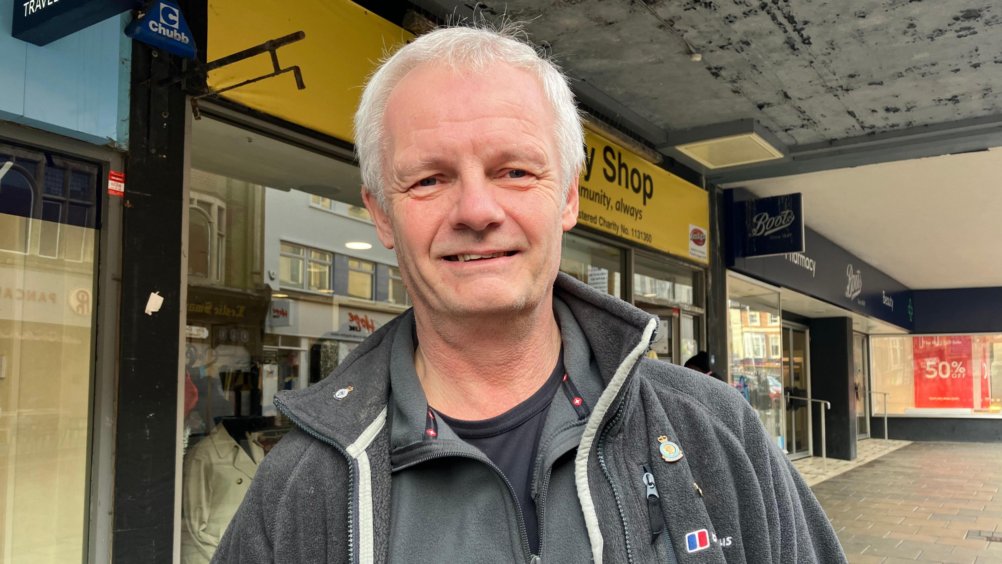 Michael Hilderley smiles at the camera as he stands outside a shop front on a pedestrianised street. He is wearing a grey fleece, grey jumper and black T-shirt. He has grey hair. 