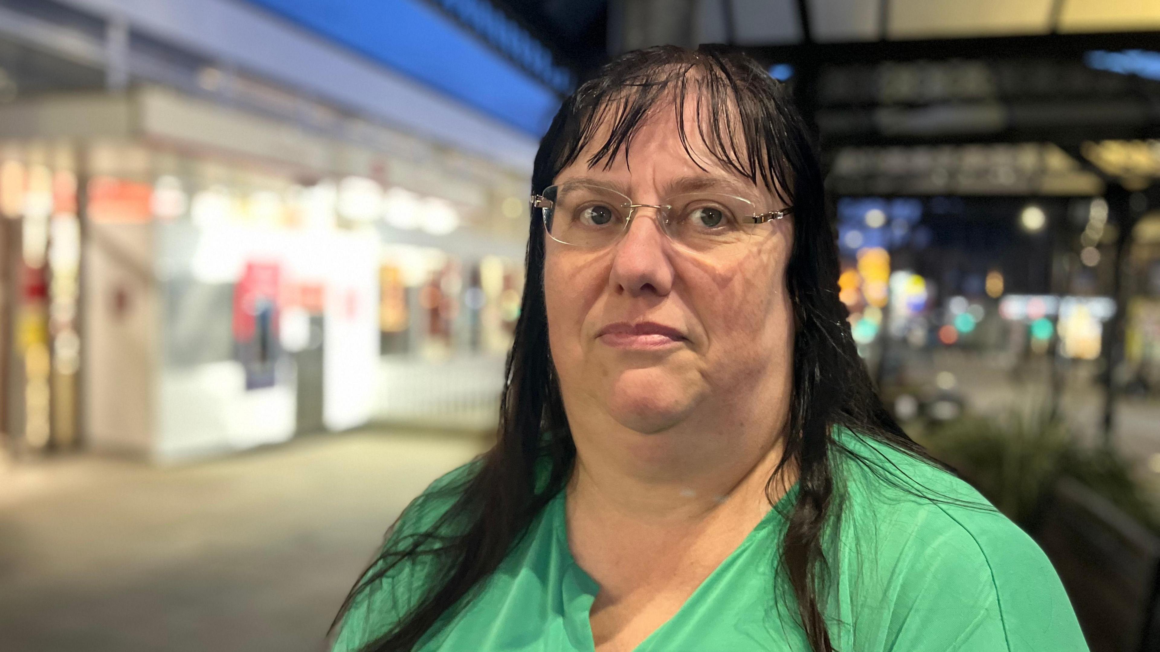 Ms Dominic wears glasses and a green blouse. She has long black hair and a fringe. The post office can be made out in the background.