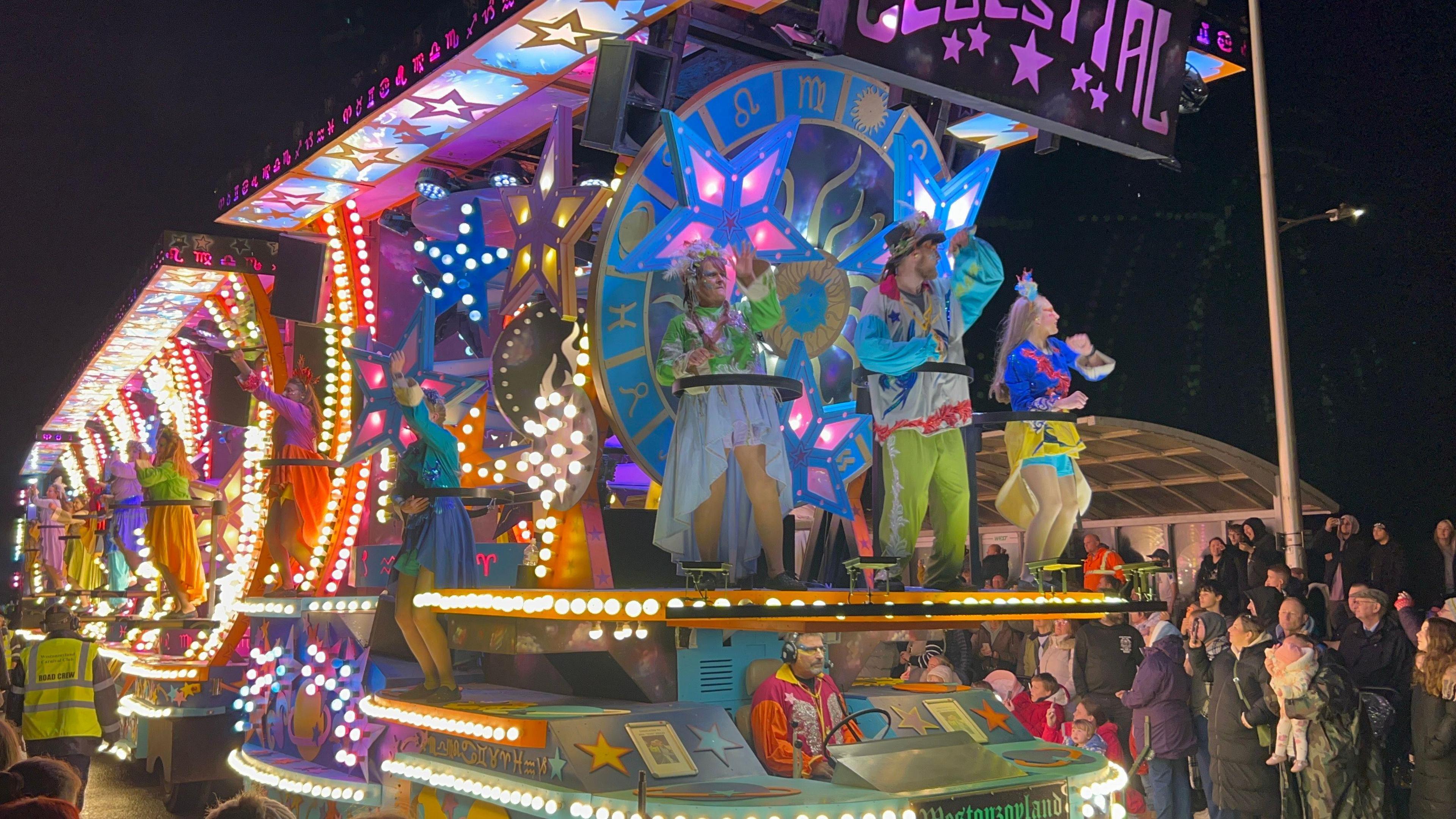 A multicoloured illuminated carnival cart. There are three performers in costumes at the front of the cart and a line of others along the side. A crowd of people can be seen watching it.