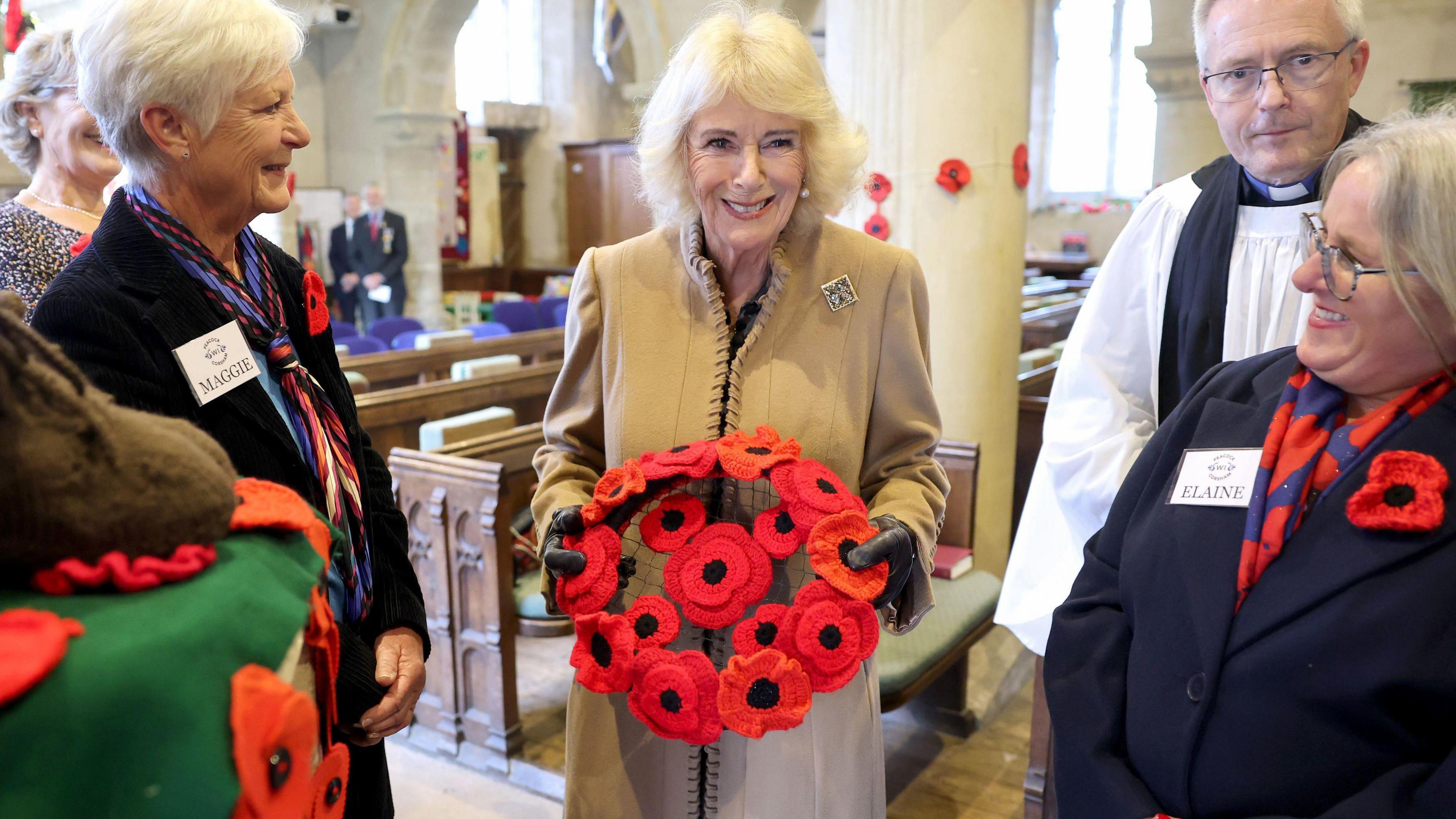 Queen Camilla holds a wreath inside St. Bartholomew's Church  surrounded by a vicar in white gown and two older ladies.