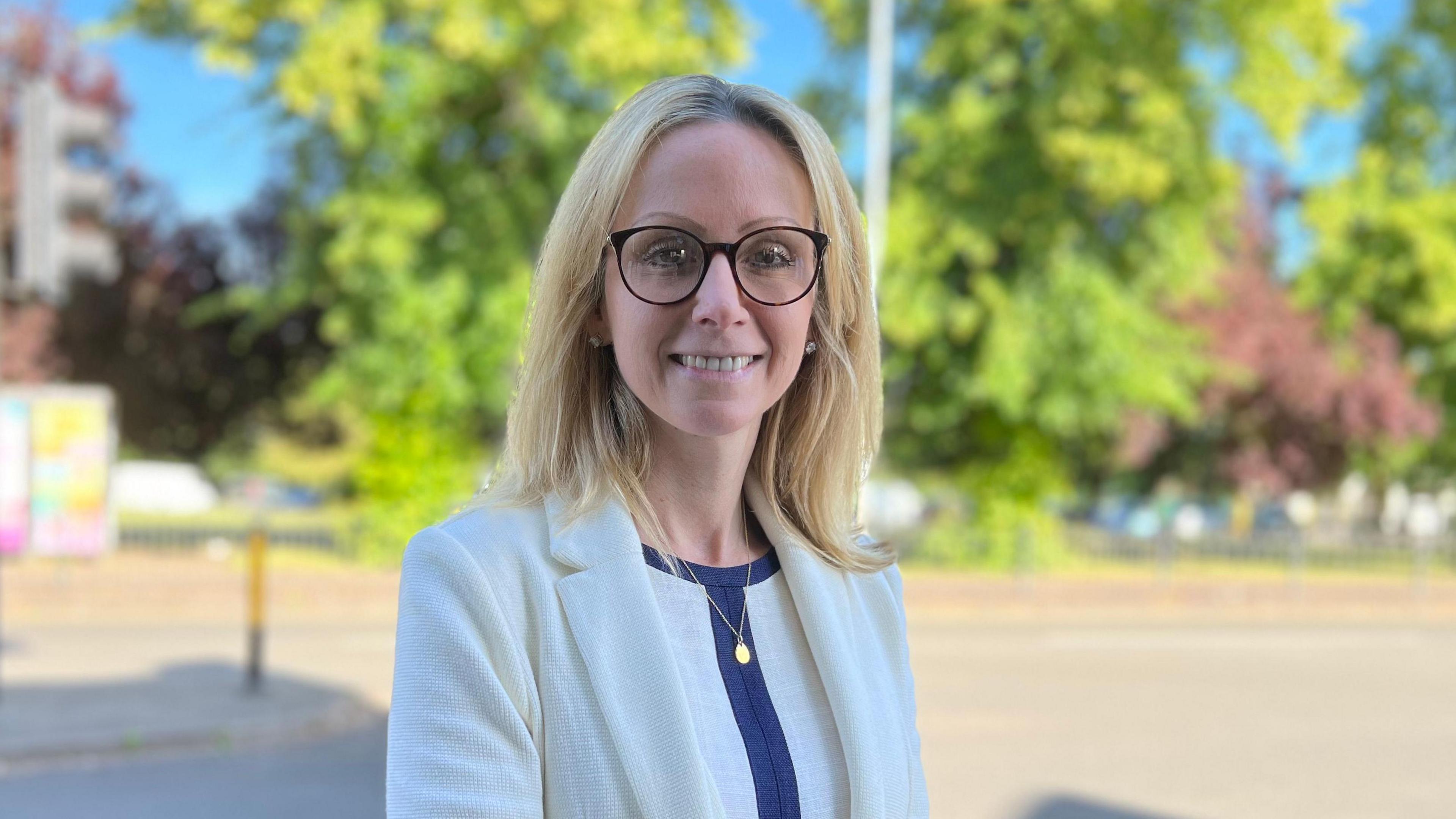 Lucy Rigby wearing a white blazer and gold necklace. She is wearing glasses and is standing against a blurred background which shows a road crossing and green trees