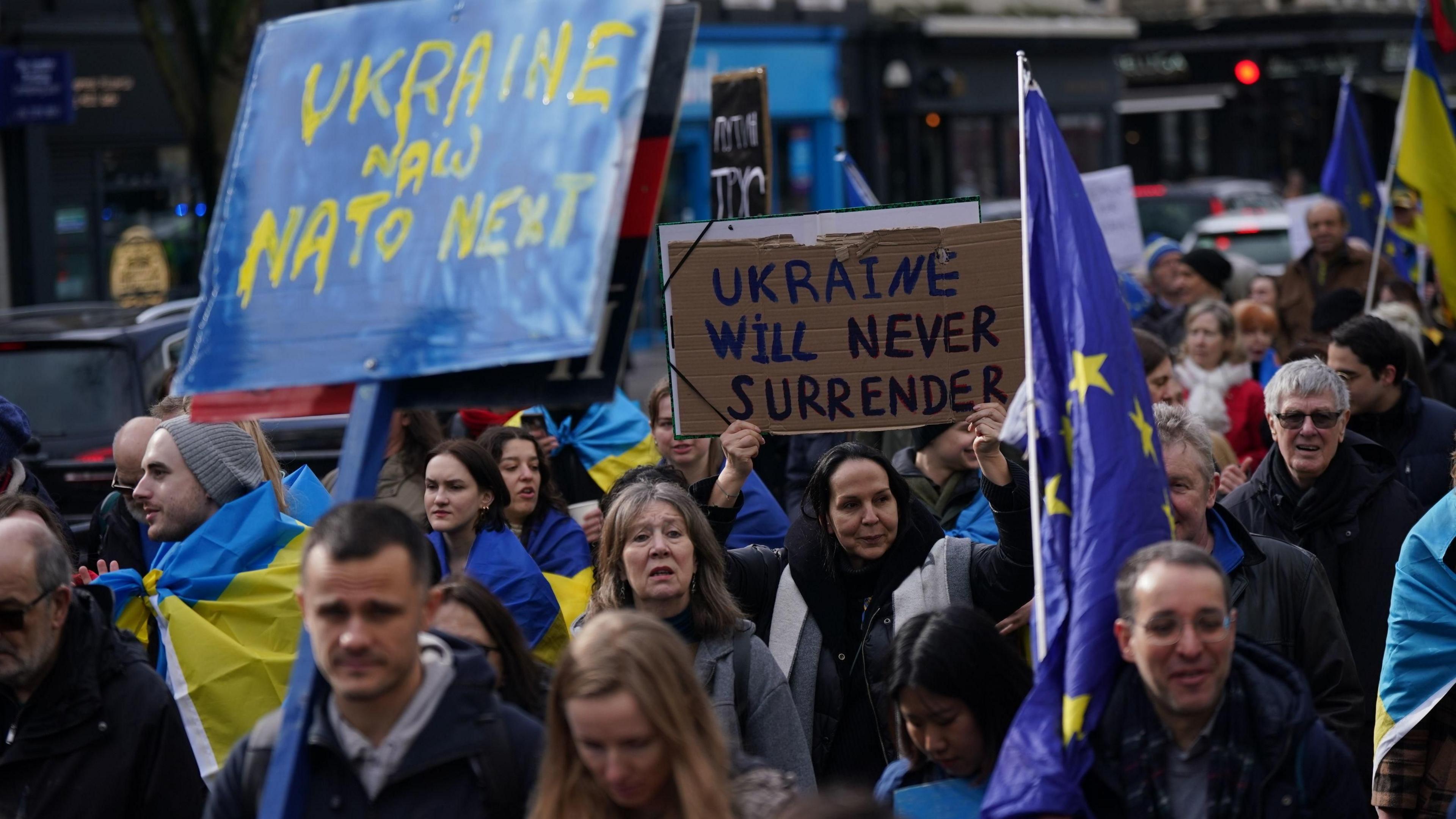 Protesters march in London, some wrapped in Ukrainian flags. One is holding an EU flag while two posters are legible, one reading: Ukraine now Nato next" and another: "Ukraine will never surrender".