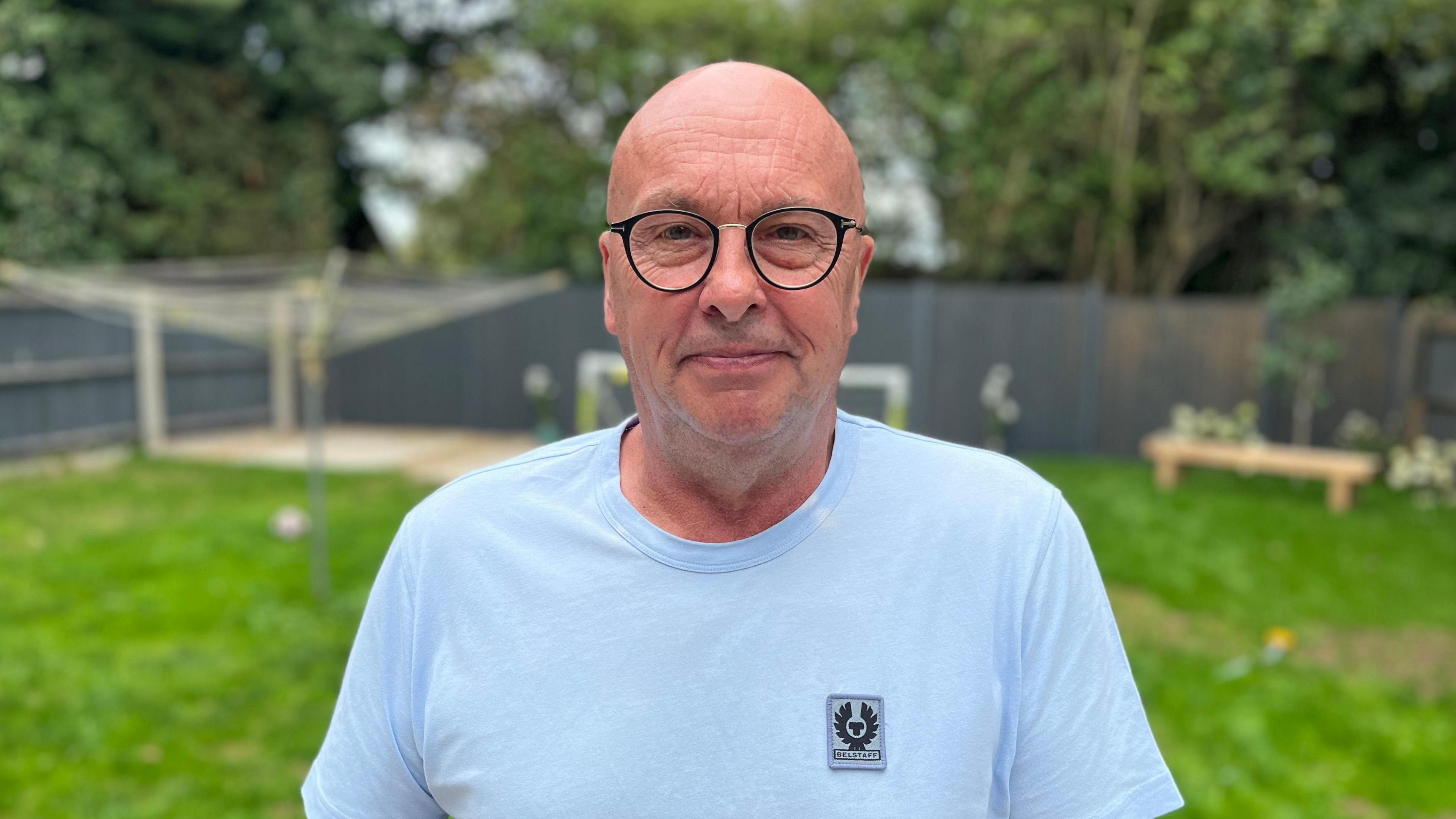 A man with glasses wearing a blue T-shirt faces the camera with a garden in the background.