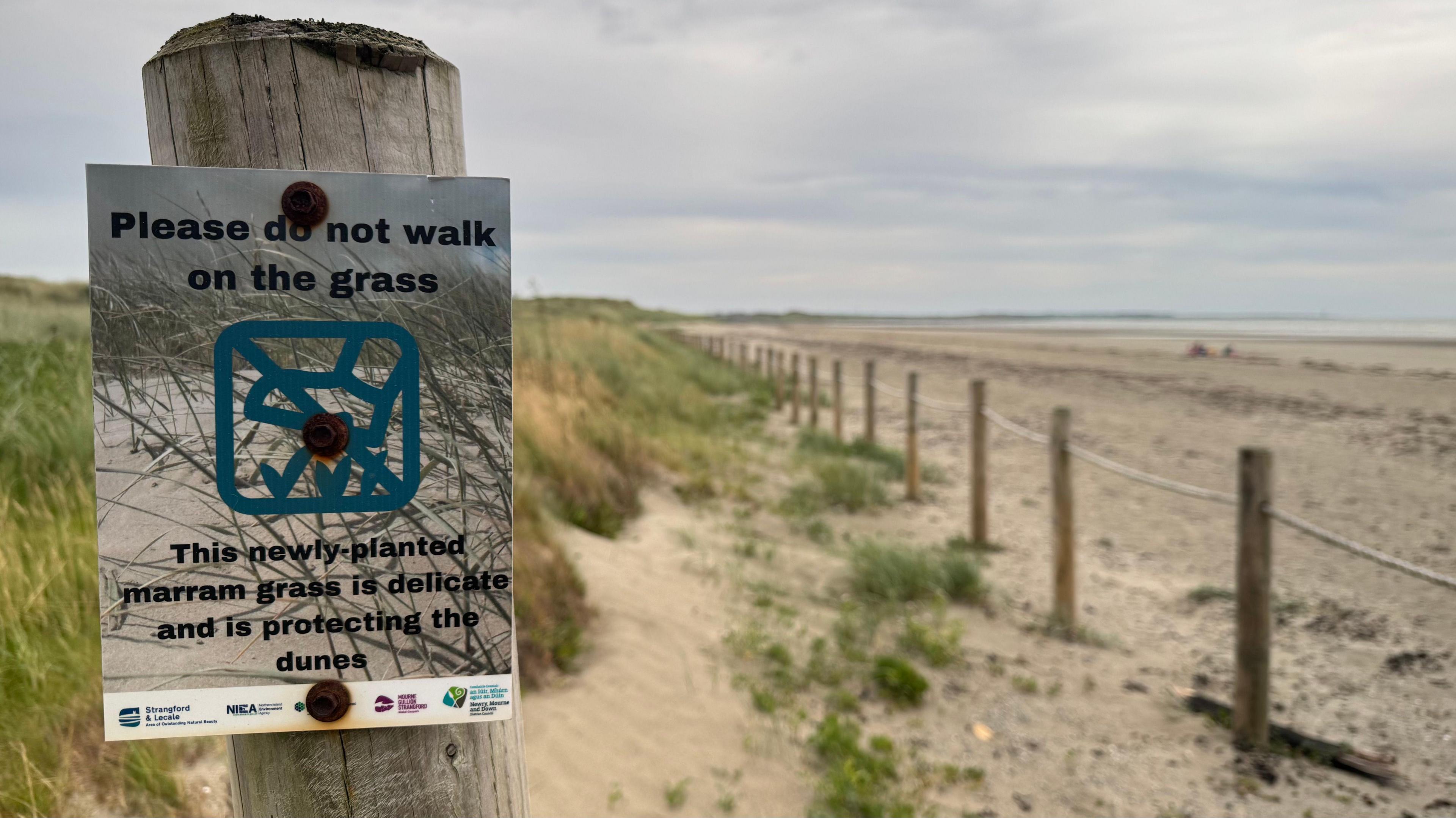 A 'please do not walk on the grass' sign is pinned to a wooden post. There is a fence and a grass in the sand dunes.