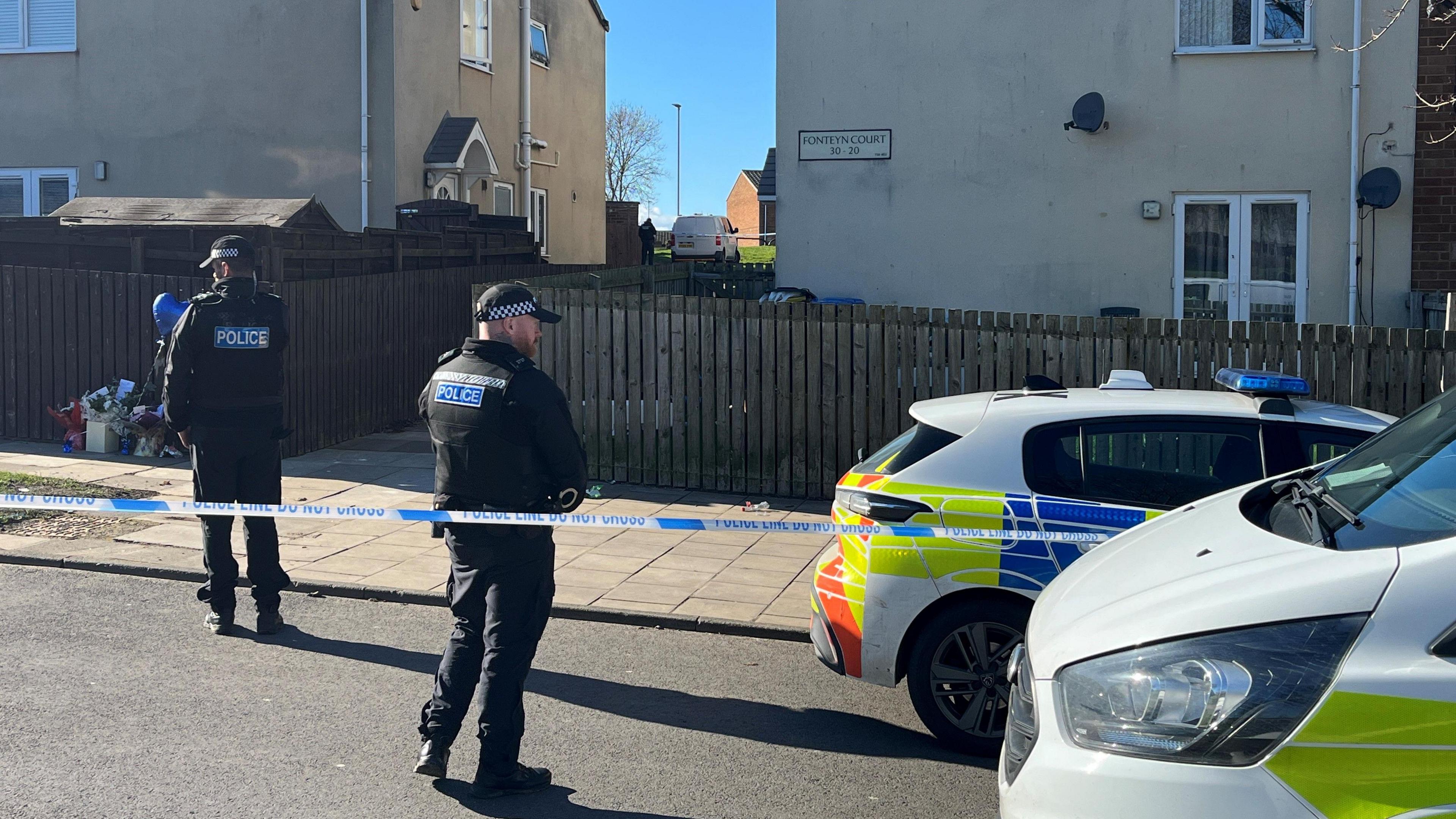 Two police officers standing behind police tape in Fonteyn Court. The tape is around an alley between two rows of houses. To the left, flowers and a balloon have been have been left next to a fence. A police car and a police van are in the foreground to the right.