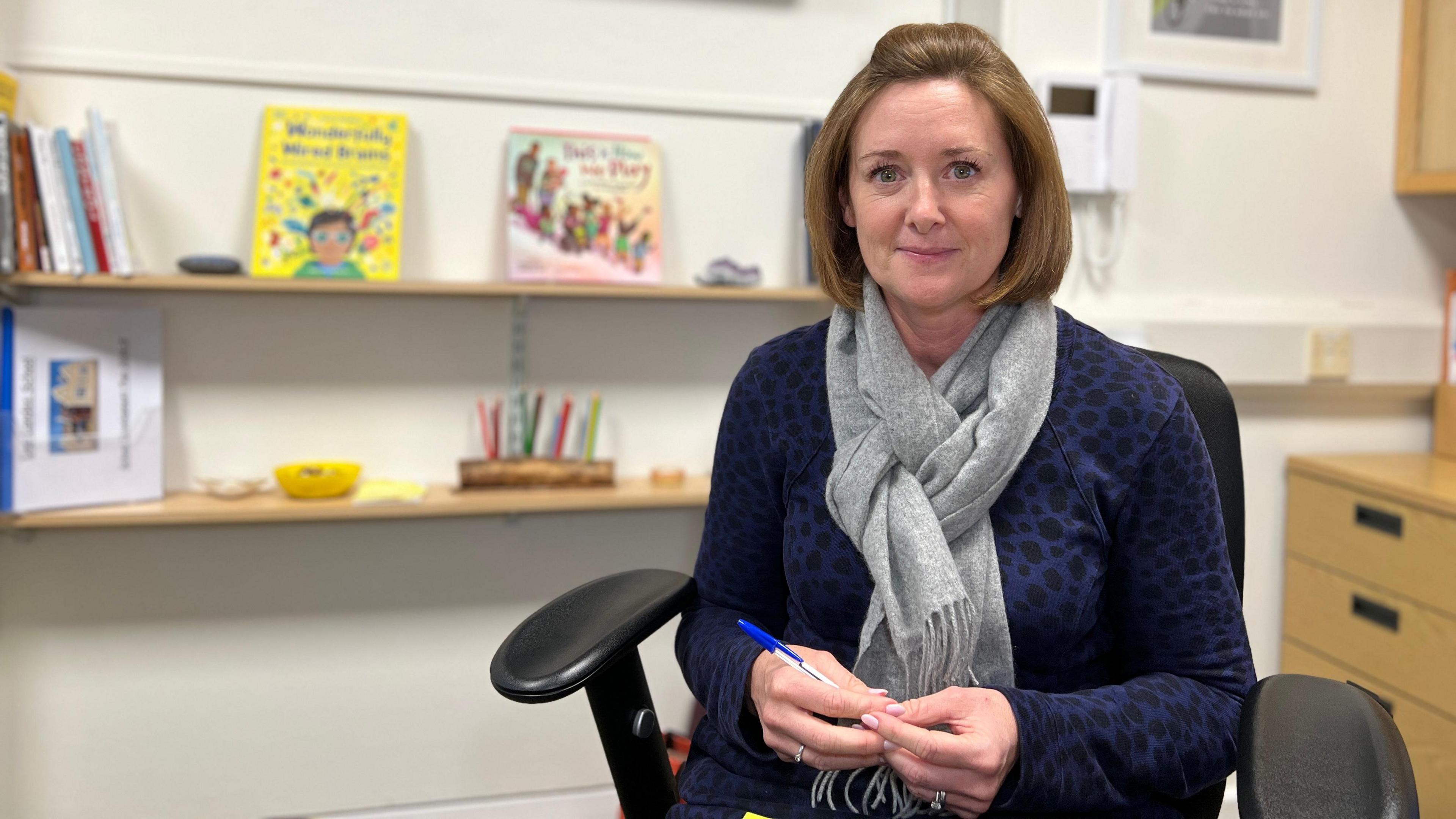 Vicki is smiling at the camera as she sits in a black chair by her desk. Behind are a few children's books on shelves. She has a dark blue with black dots jumper and a grey scarf. She's holding a blue biro pen in her hands and has short brown hair.