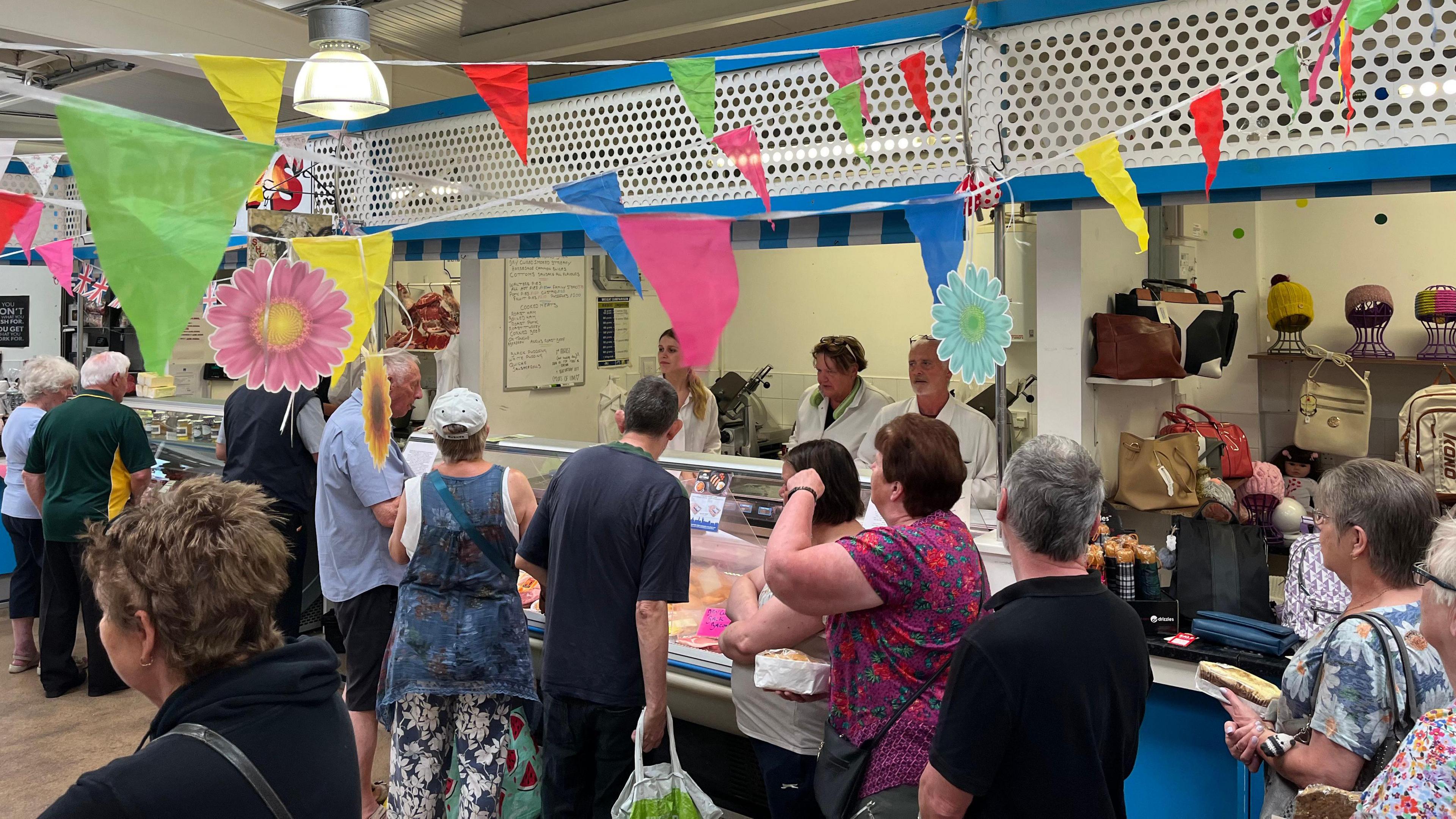 Multiple customers queue outside a meat market, where three members of staff stand in white shirts behind the counter, with colourful bunting above them