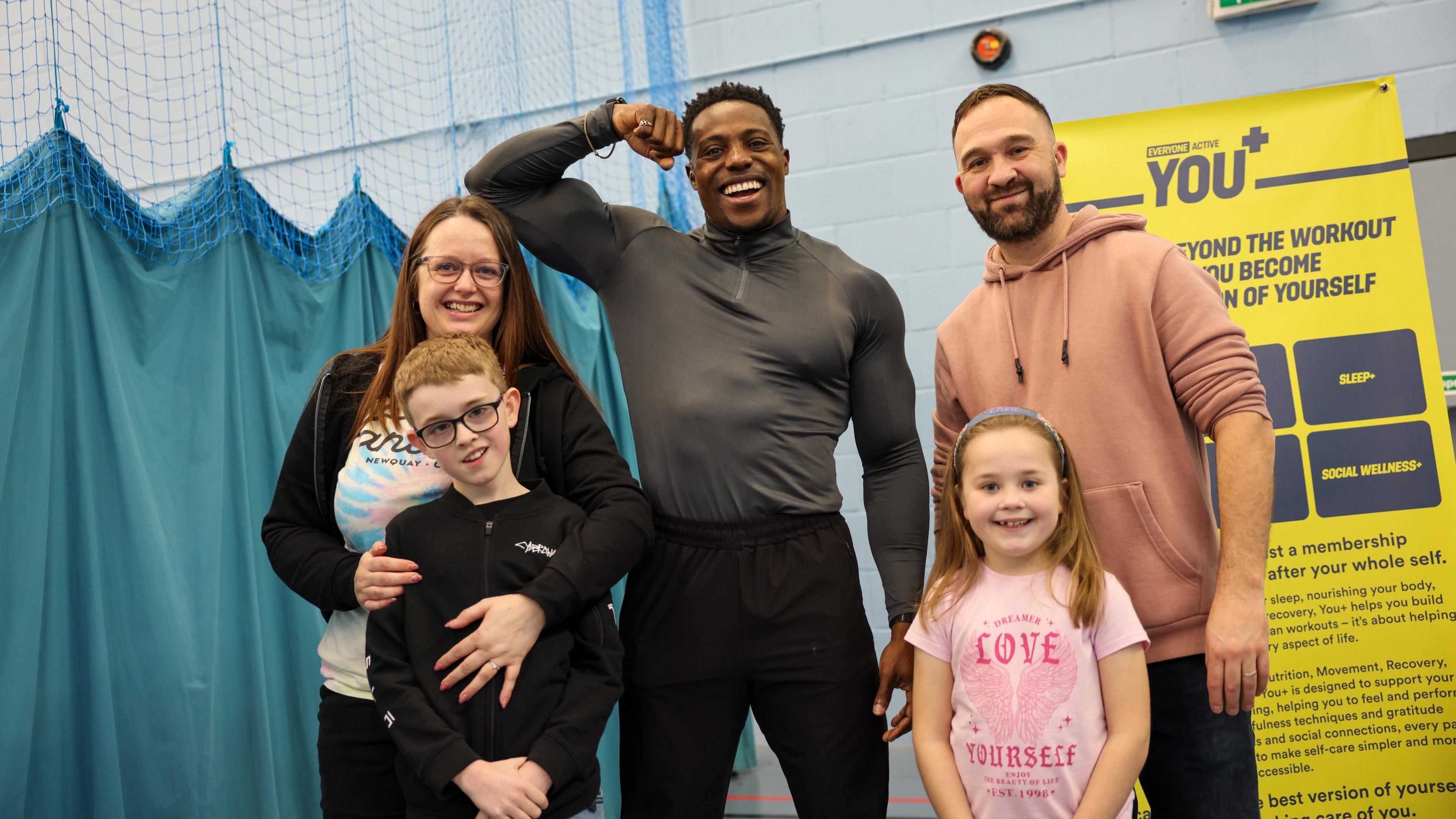 Harry Aikines-Aryeetey wearing black tracksuit bottoms and a grey long-sleeved gym shirt. He is posing with his bicep flexed and smiling at the camera. Beside him are a family with two small children, who are all smiling at the camera. They are standing in the Horfield Leisure Centre gym in front of a blue net curtain.