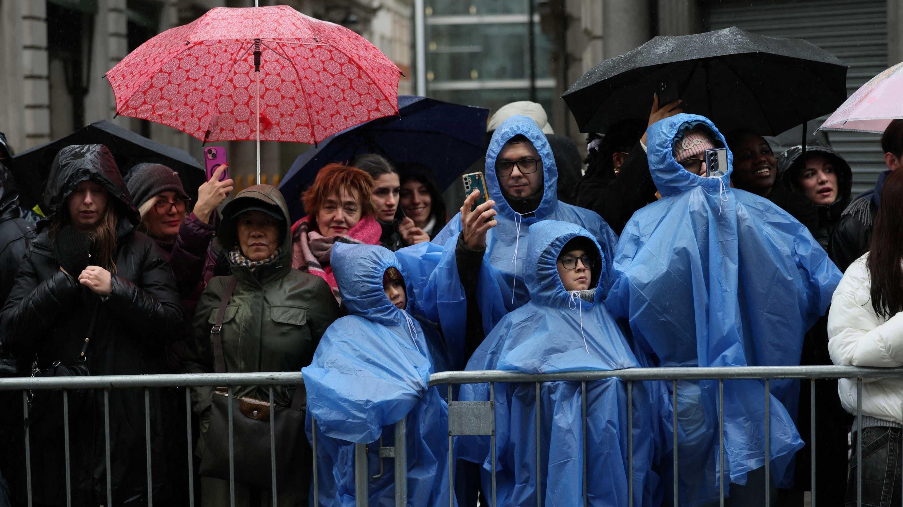 Two adults and two children wearing blue rain ponchos stand among a crown of people holding umbrellas behind crowd barriers. 