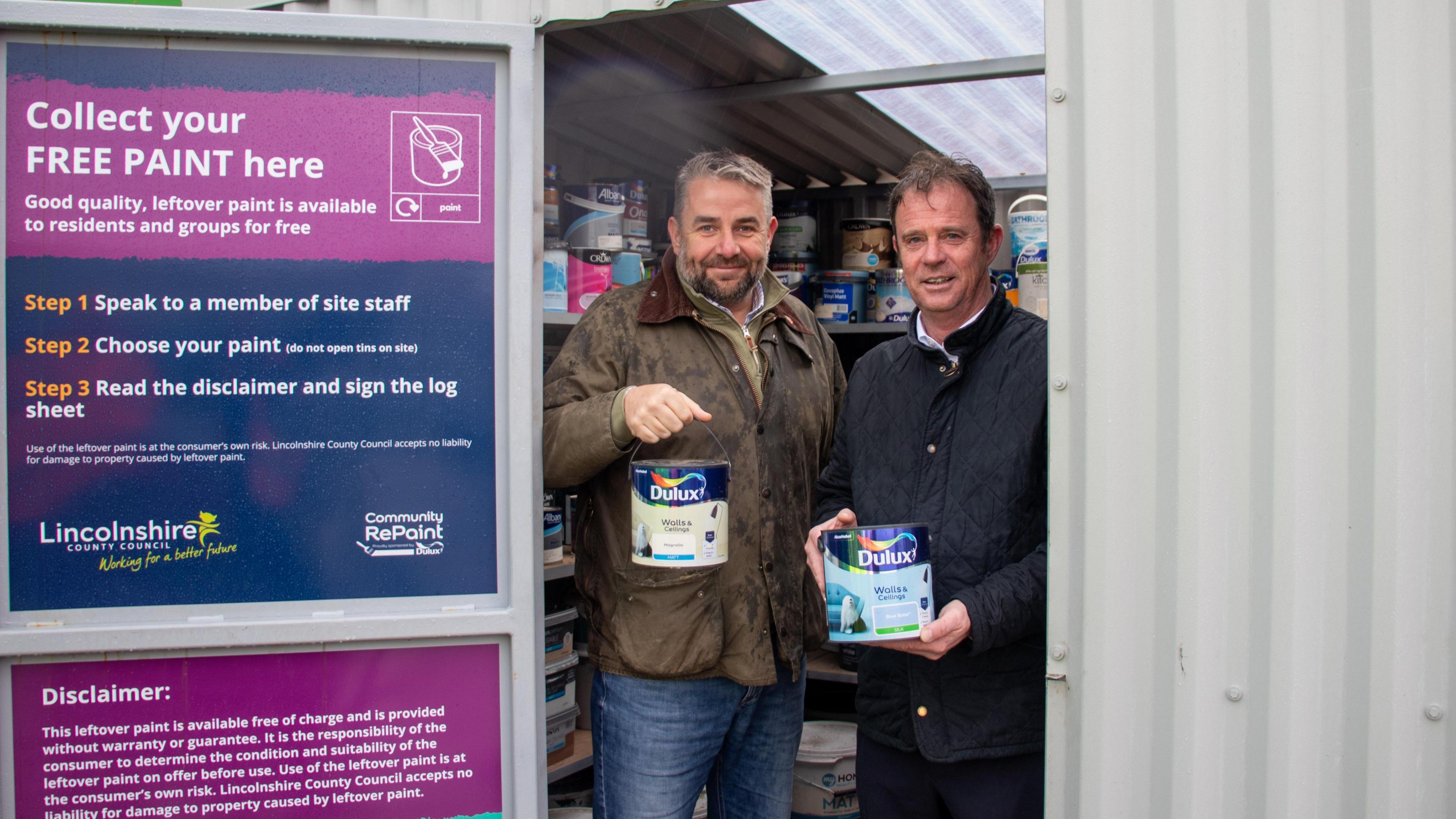 Two men stand inside a corrugated metal shed holding paint tins next to a sign for a new paint recycling scheme.