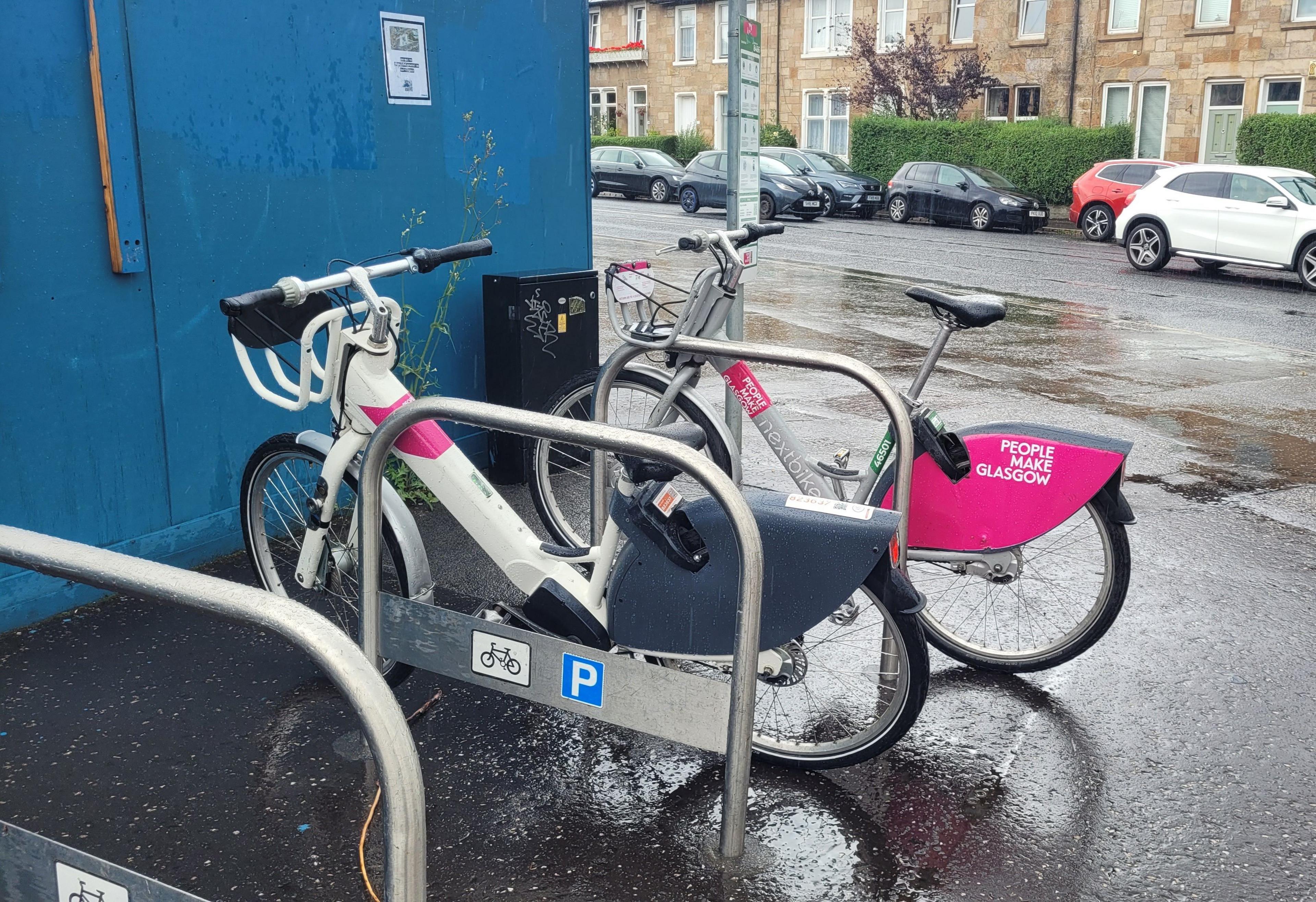 Two nextbikes sit in a bike rack in Glasgow. ONe is electric and one is non-electric. The non-electric bike has "people make Glasgow" written on it.