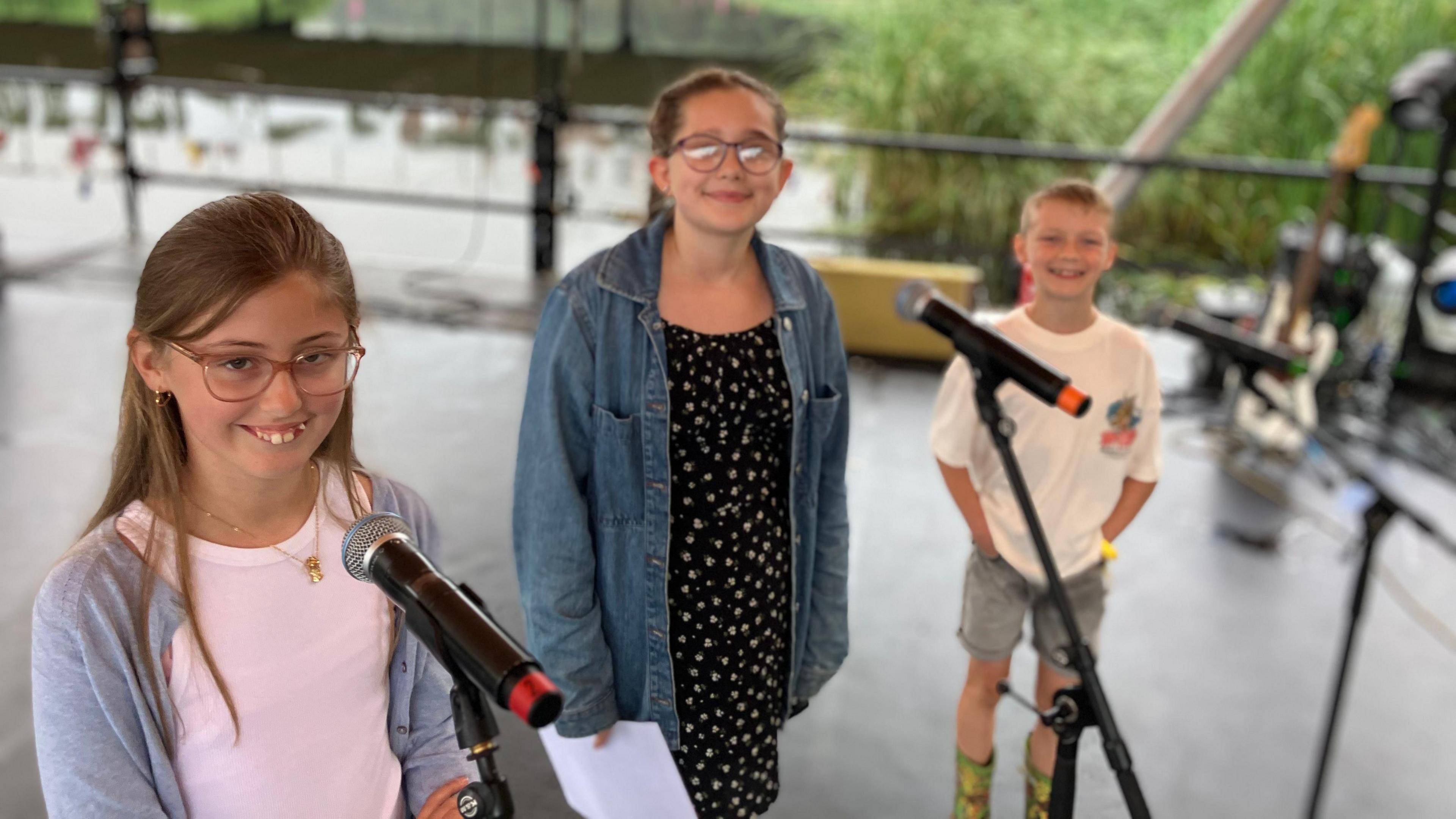 Two 11 year old girls and a 7 year old boy stage on a festival stage with microphones in front of the,