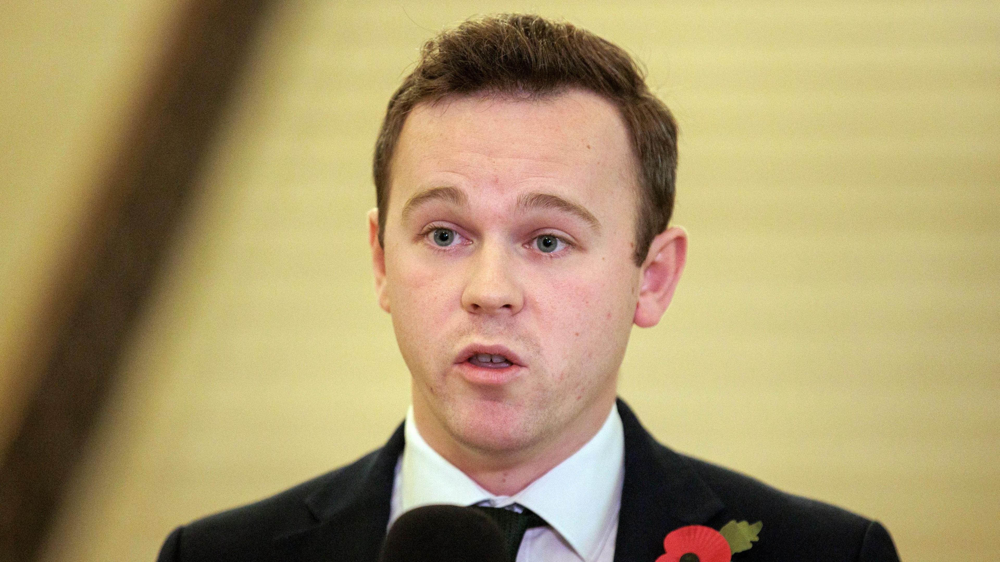 Eóin Tennyson, a man with short dark brown hair, in a black suit, pictured speaking to reporters. He has a poppy on his lapel.
