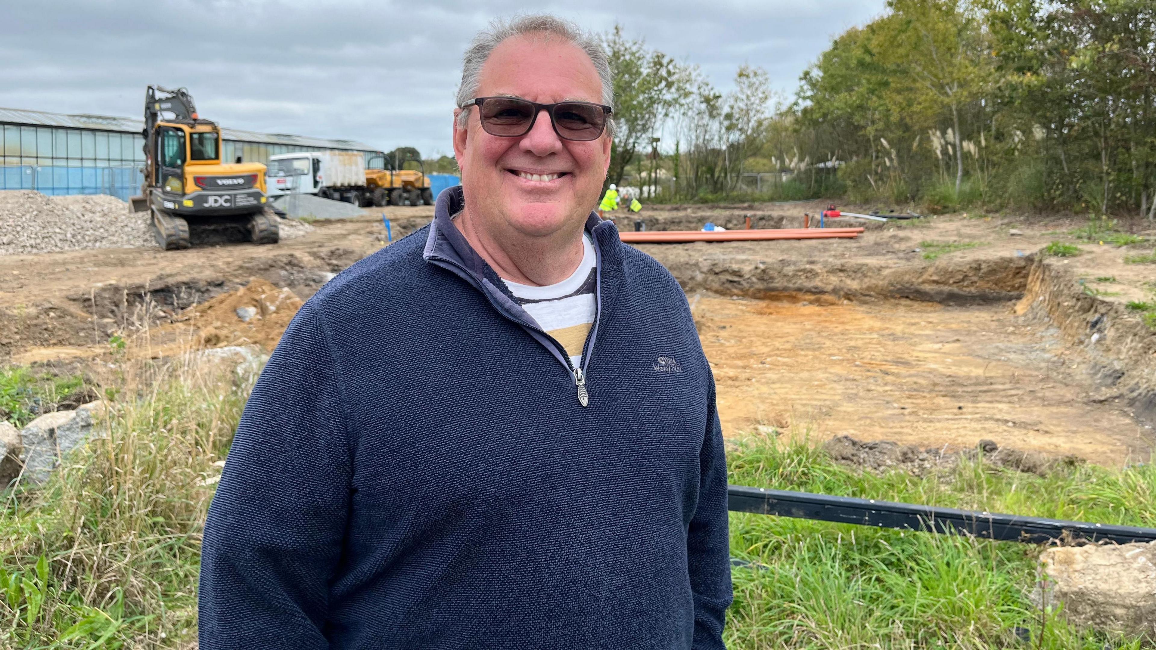A smiling man in a blue fleece and sunglasses stands in front of a building site where grass has been removed. A digger is in the background.