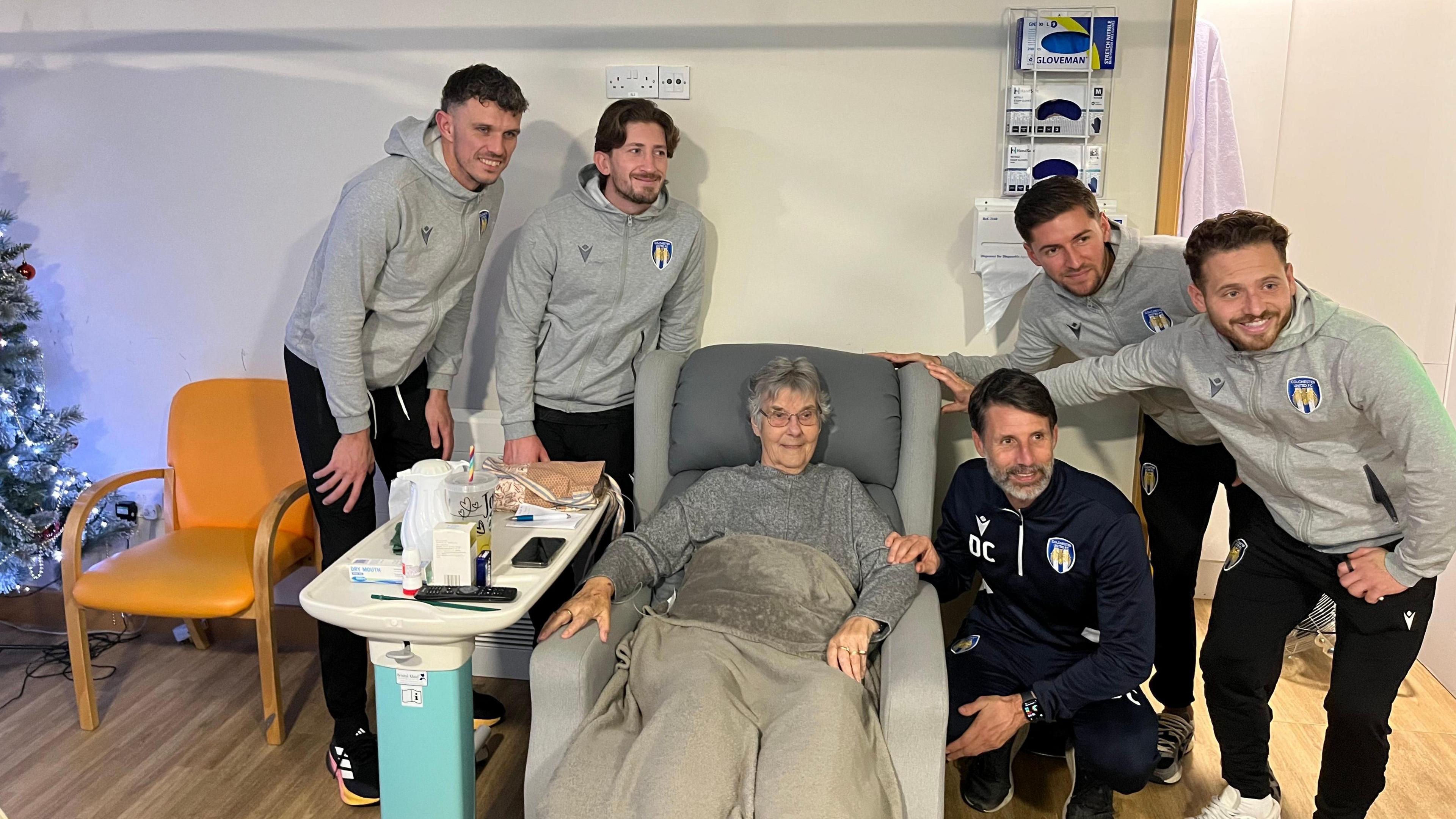 Footballers wearing grey hoodies standing by an elderly woman who is sitting on a sofa with a blanket. They are all smiling at the camera.