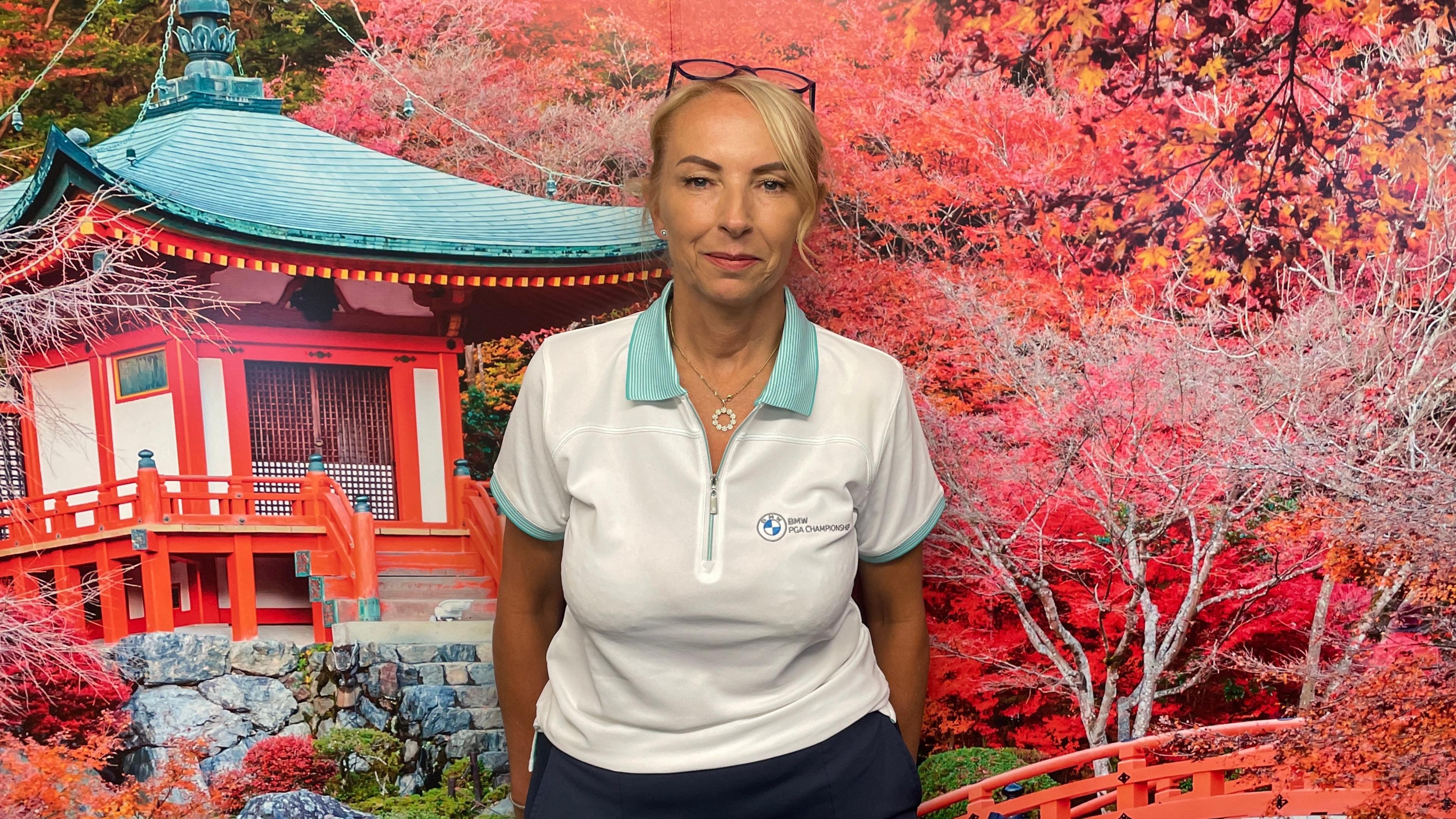 Jules Armstrong standing in front of a wall decorated with an autumnal scene of flowers and trees outside a house. She has blonde hair, glasses balanced on her head and wears a white polo shirt with green edging