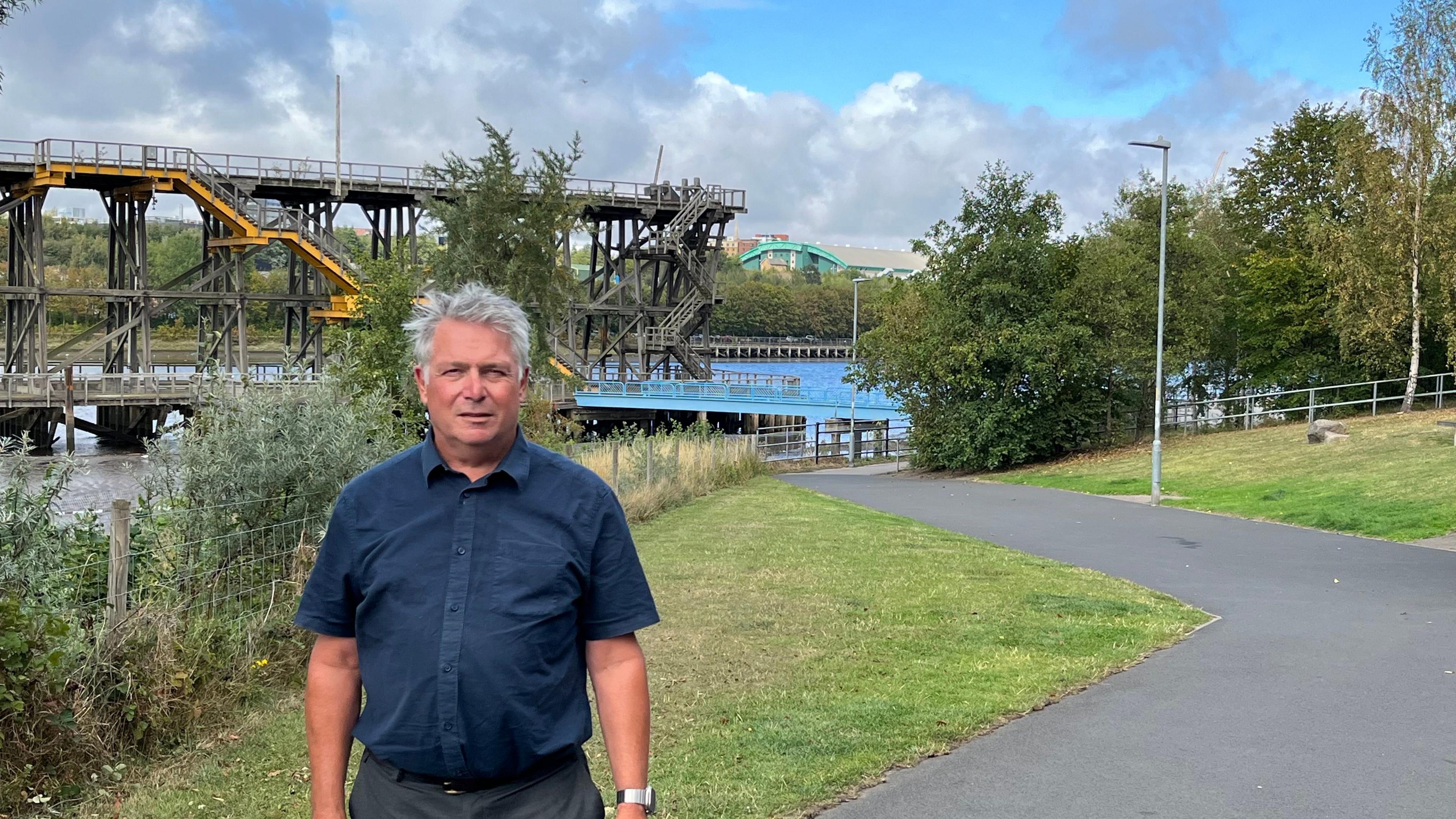 Councillor Gary Haley on the Riverside Row path. He has grey hair and is wearing a navy short-sleeved shirt. Grass and trees are either side of the path, while Dunston Staiths - a wooden structure once used to load coal on to ships on the river - can be seen in the background.